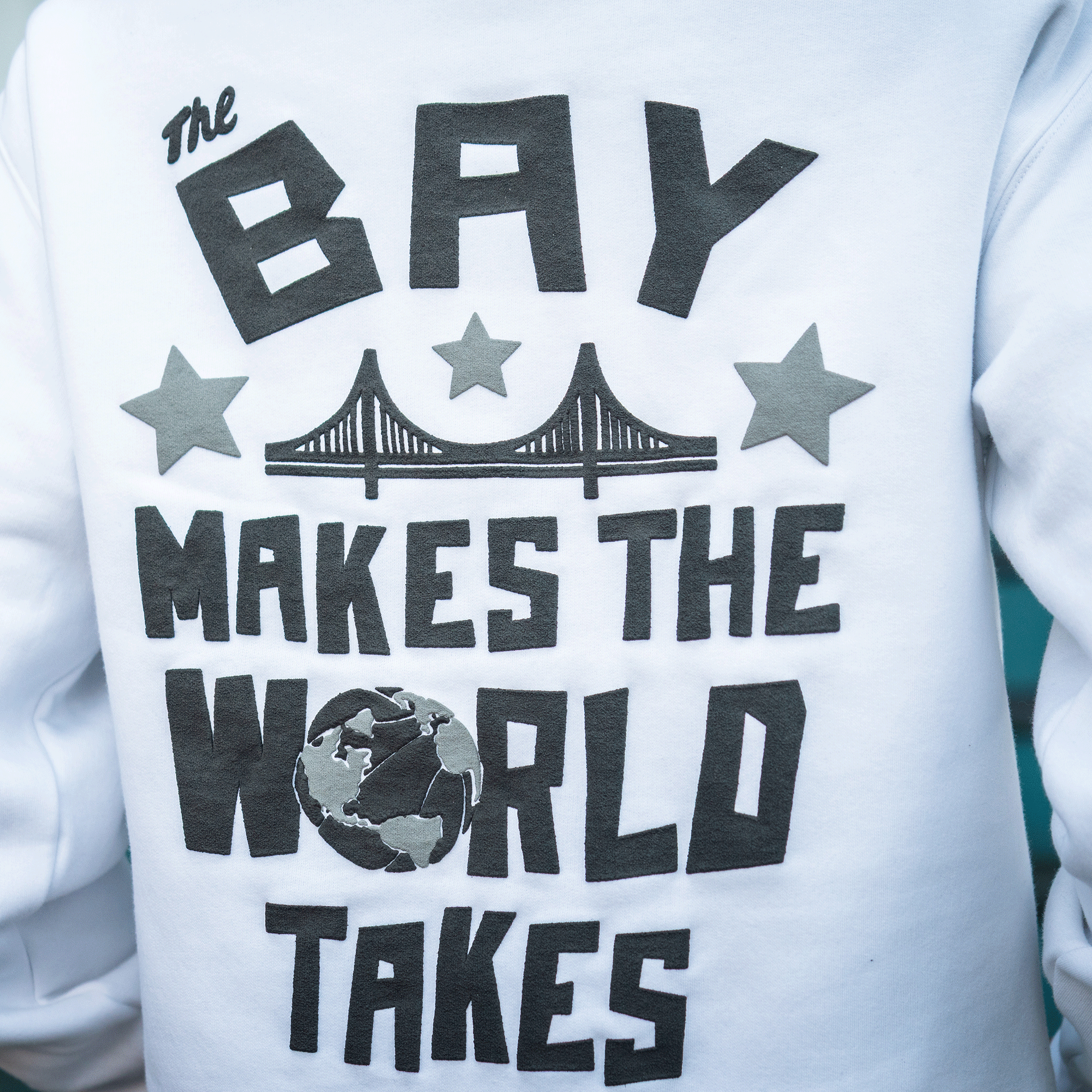 The Bay Bridge Hoodie By DOC V2 from Oaklandish features bold “The Bay Makes The World Takes” text with two stars, a Bay Bridge graphic, and an Earth image as the "O" in “World.”.
