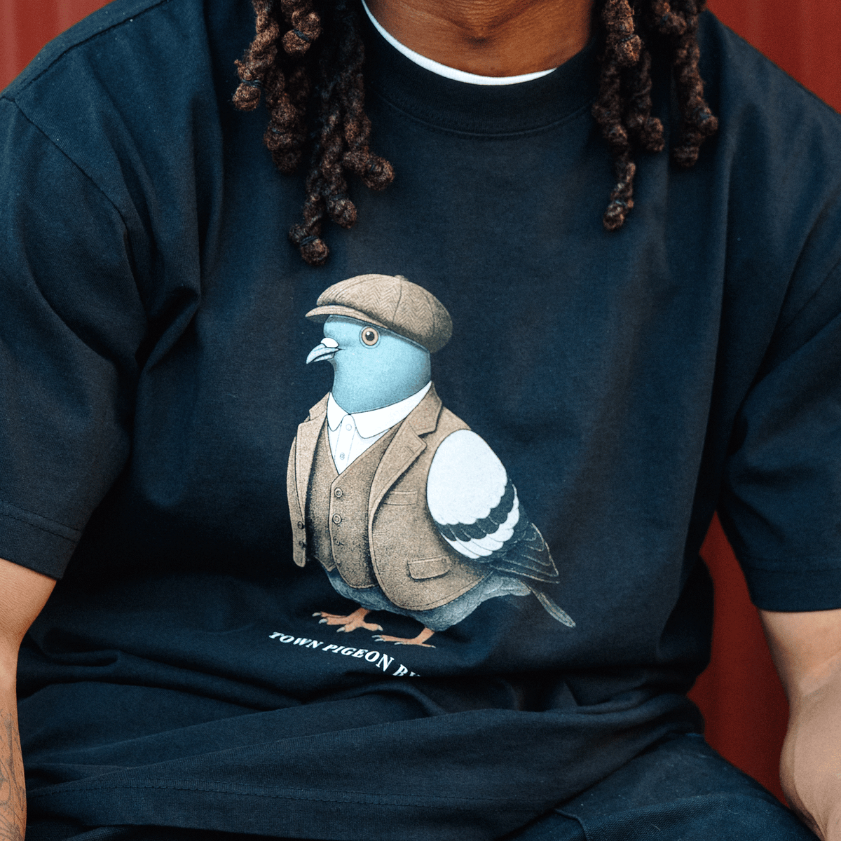 A person wears an Oaklandish Dapper Tee, a heavy-weight cotton crew-neck t-shirt featuring a centered graphic of a pigeon dressed in a beige tweed vest, white collared shirt, and matching flat cap, with small, partially obscured text printed below the dapper bird.