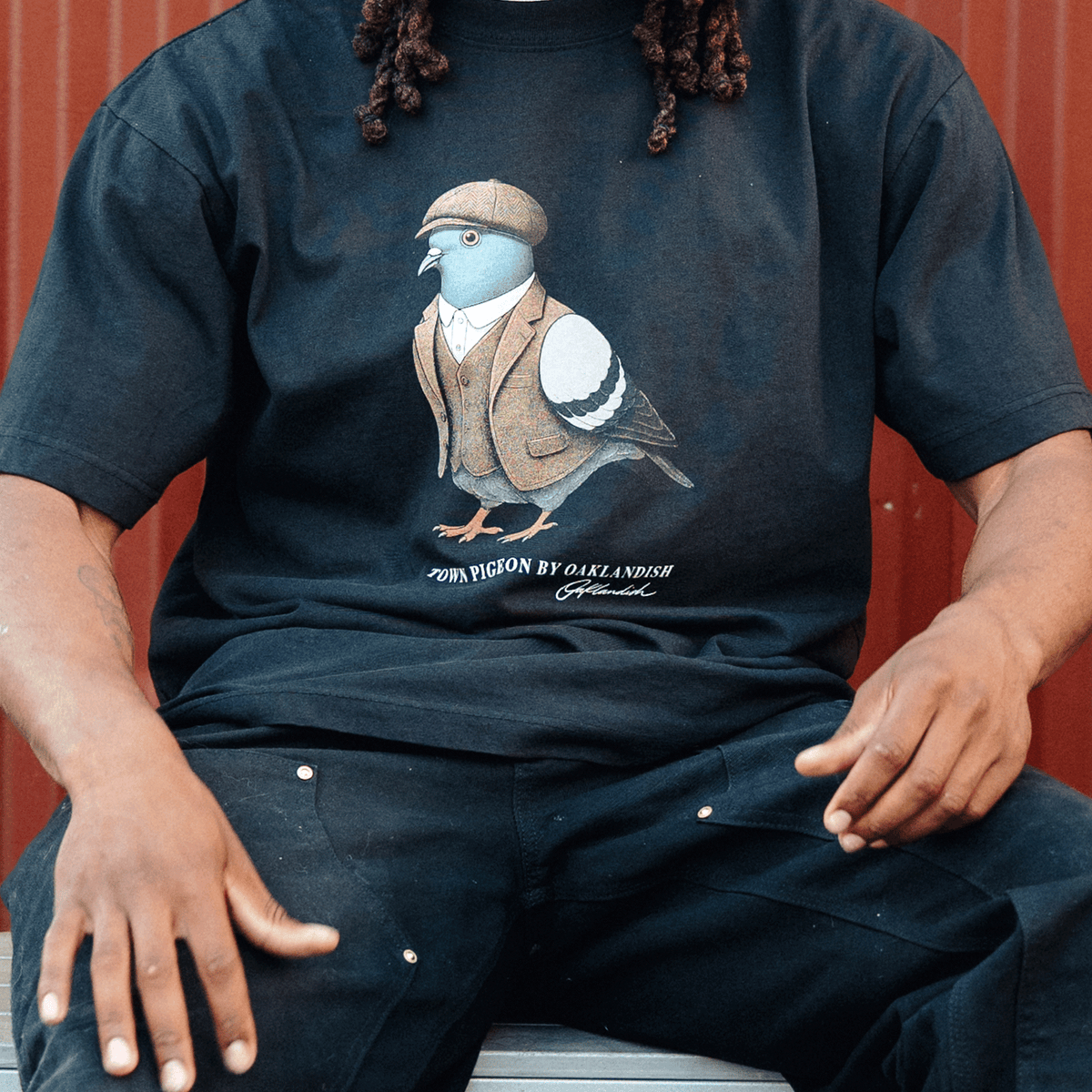 A person wears a black, heavy-weight cotton Dapper Tee featuring a stylized illustration of a pigeon in a tweed vest and flat cap, with “TOWN PIGEON” in white letters against a deep red background.