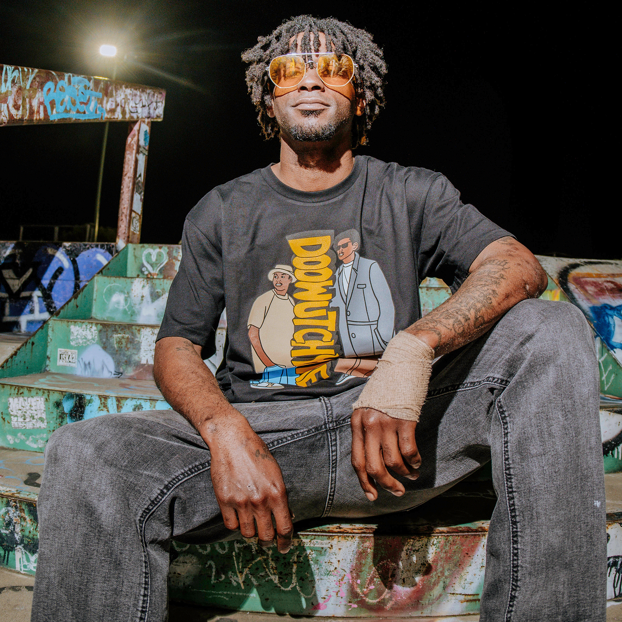 A man with dreadlocks and yellow sunglasses sits on graffiti-covered steps at night, exuding 90s vibes. He wears a Doowutchyalike Tee by Oaklandish, gray jeans, and a hand wrap. Under the streetlight and cartoon-style backdrop, he smiles while leaning back, utterly relaxed.
