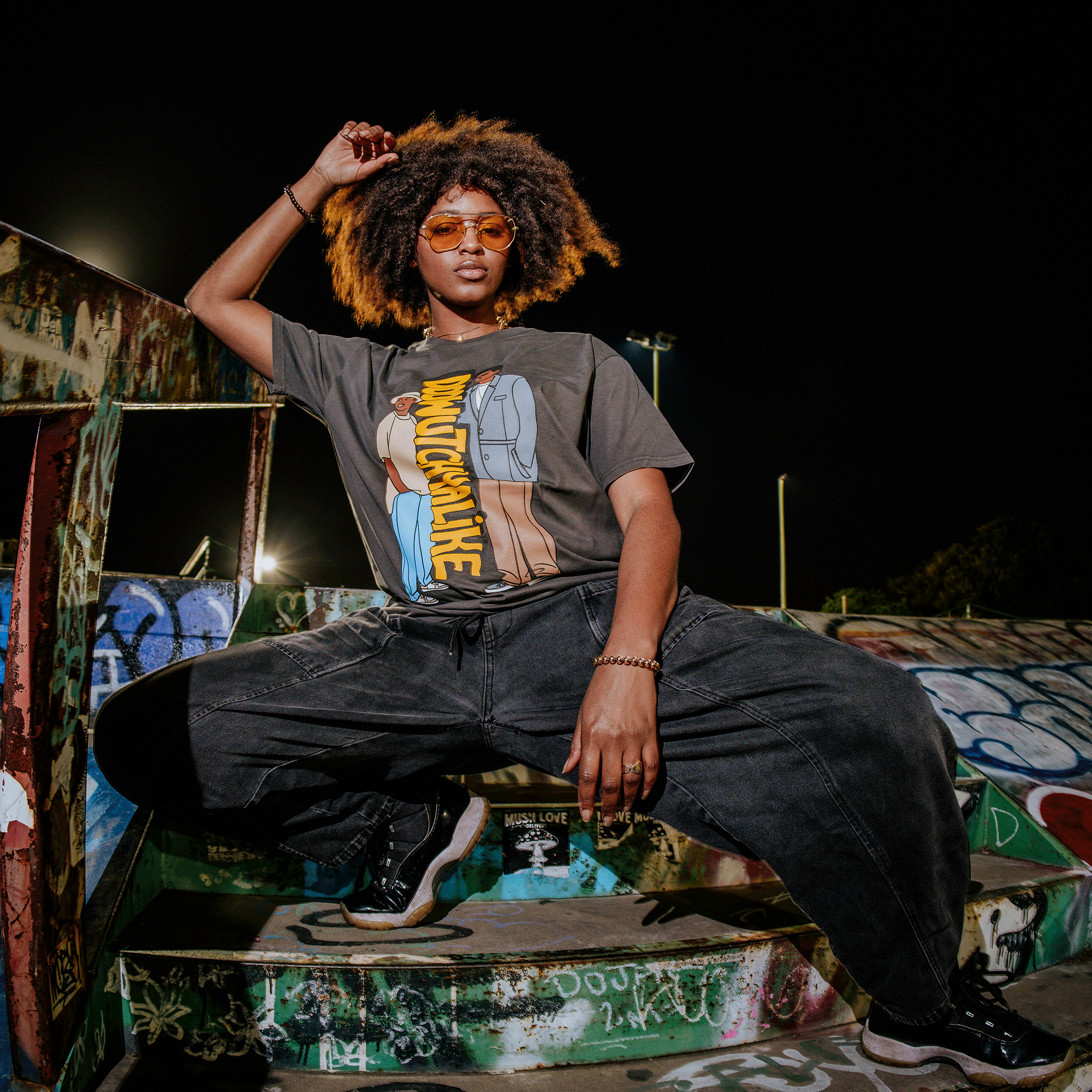 In a graffiti-covered skatepark, a person with curly hair and sunglasses confidently poses 90s-style. Wearing Oaklandish's Doowutchyalike Tee and baggy pants, they squat on the spray-painted ramp, surrounded by vibrant artwork that captures Digital Underground vibes.