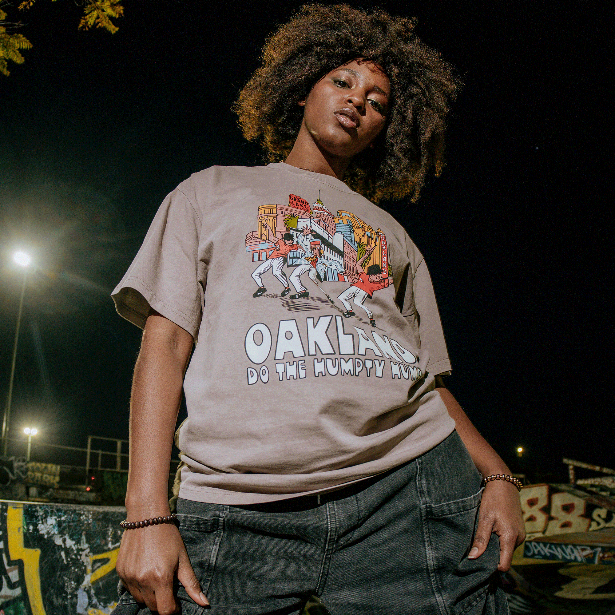 Under streetlights with graffiti in the background, a curly-haired person in light jeans confidently wears the "Do the Humpty Hump Heavy Tee" by Oaklandish. The shirt displays "OAKLAND DO THE HUMPTY HUMP" with vivid cartoons and a cityscape, celebrating Digital Underground.