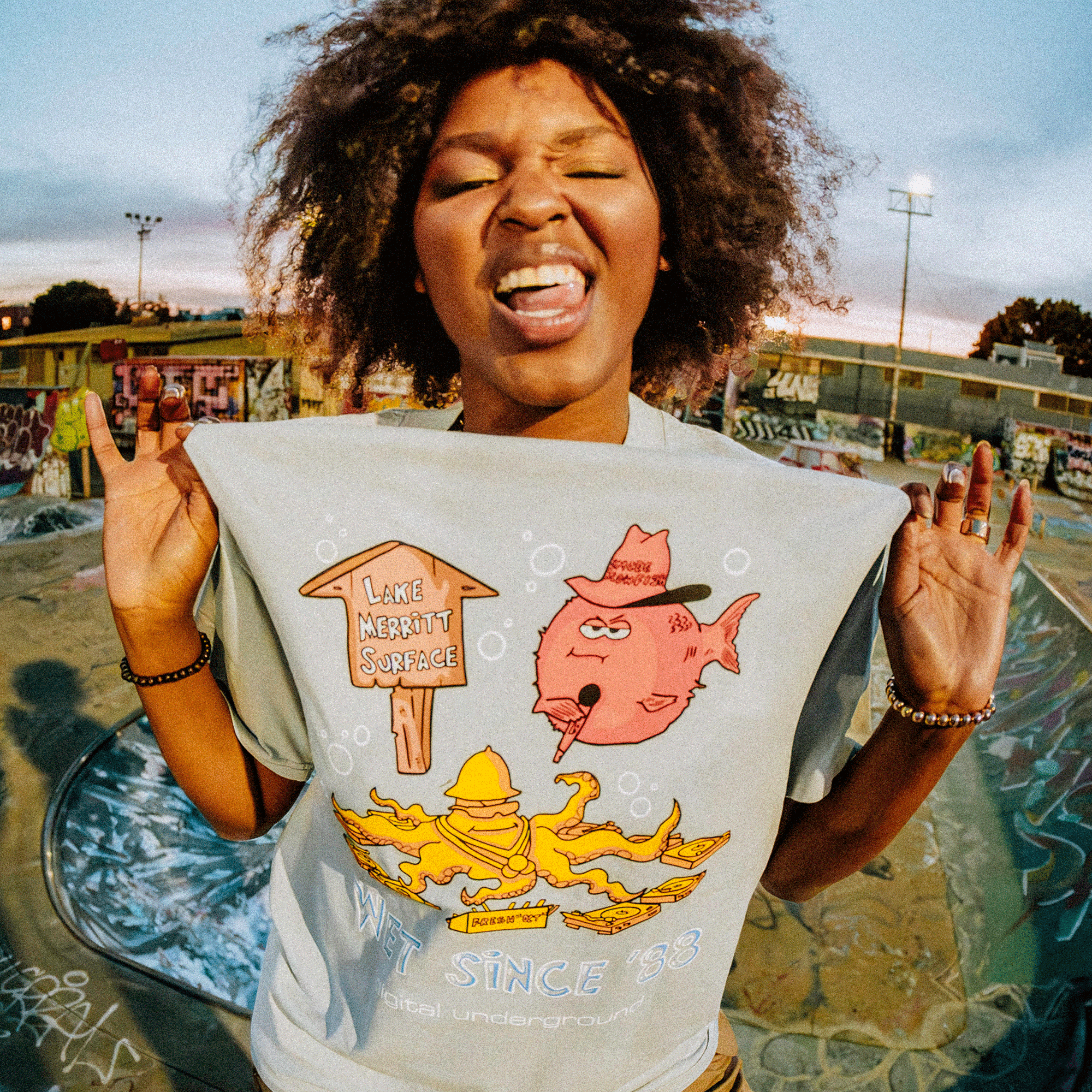 In an Oakland skatepark filled with graffiti-covered ramps, a person with curly hair playfully tugs their Wet Since '88 Heavy Tee by Oaklandish, featuring cartoons and the text "Lake Merritt Surface." They joyfully stick out their tongue and close their eyes.