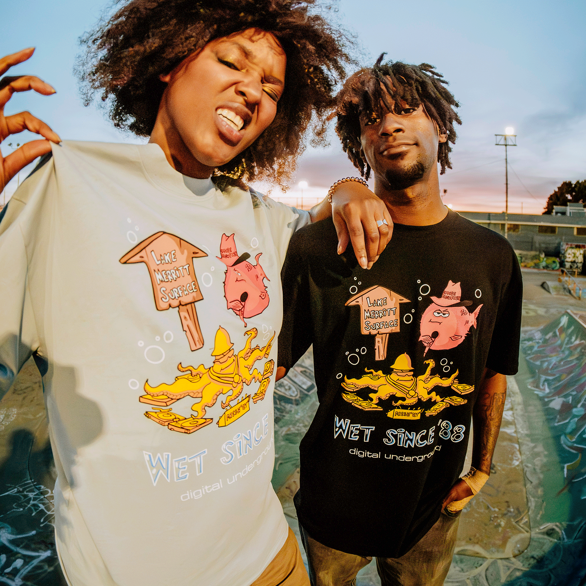 Two people are outside in Oakland wearing identical "Wet Since '88" tees by Oaklandish, featuring a playful fish design and the phrase "Like Nobody's Business." In front of a skate park, one of them humorously mimics Digital Underground's style.