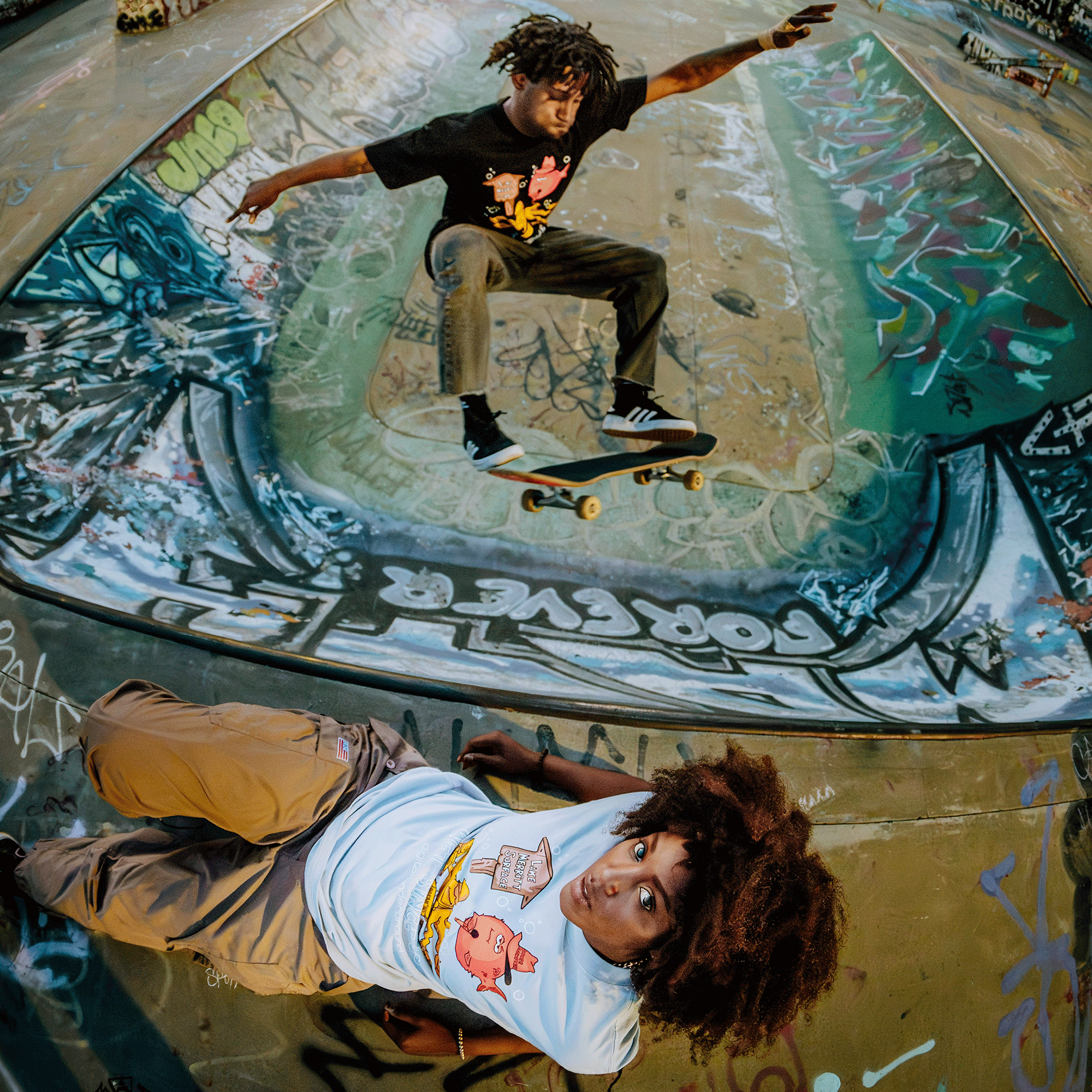 A skateboarder in a black shirt performs an aerial trick over a graffiti-covered skate bowl, echoing Oaklandish's vibrant roots. In the foreground, someone in a Wet Since '88 Heavy Tee by Oaklandish with an afro hairstyle lies on the ground, gazing at the camera—dynamic and artistic.