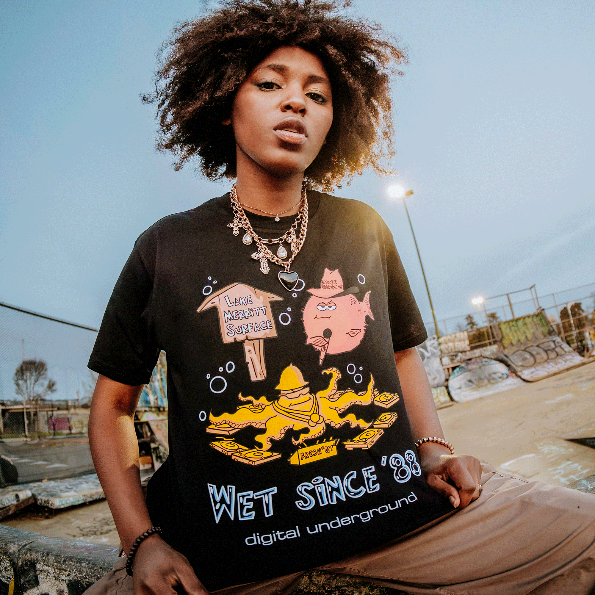 In an overcast Oakland skate park, a curly-haired individual stands confidently at dusk. They're wearing the Wet Since '88 Tee by Oaklandish, a black graphic shirt with yellow cartoon figures and the text "Wet Since '88, Digital Underground." Graffiti adorns the ramps in the background.