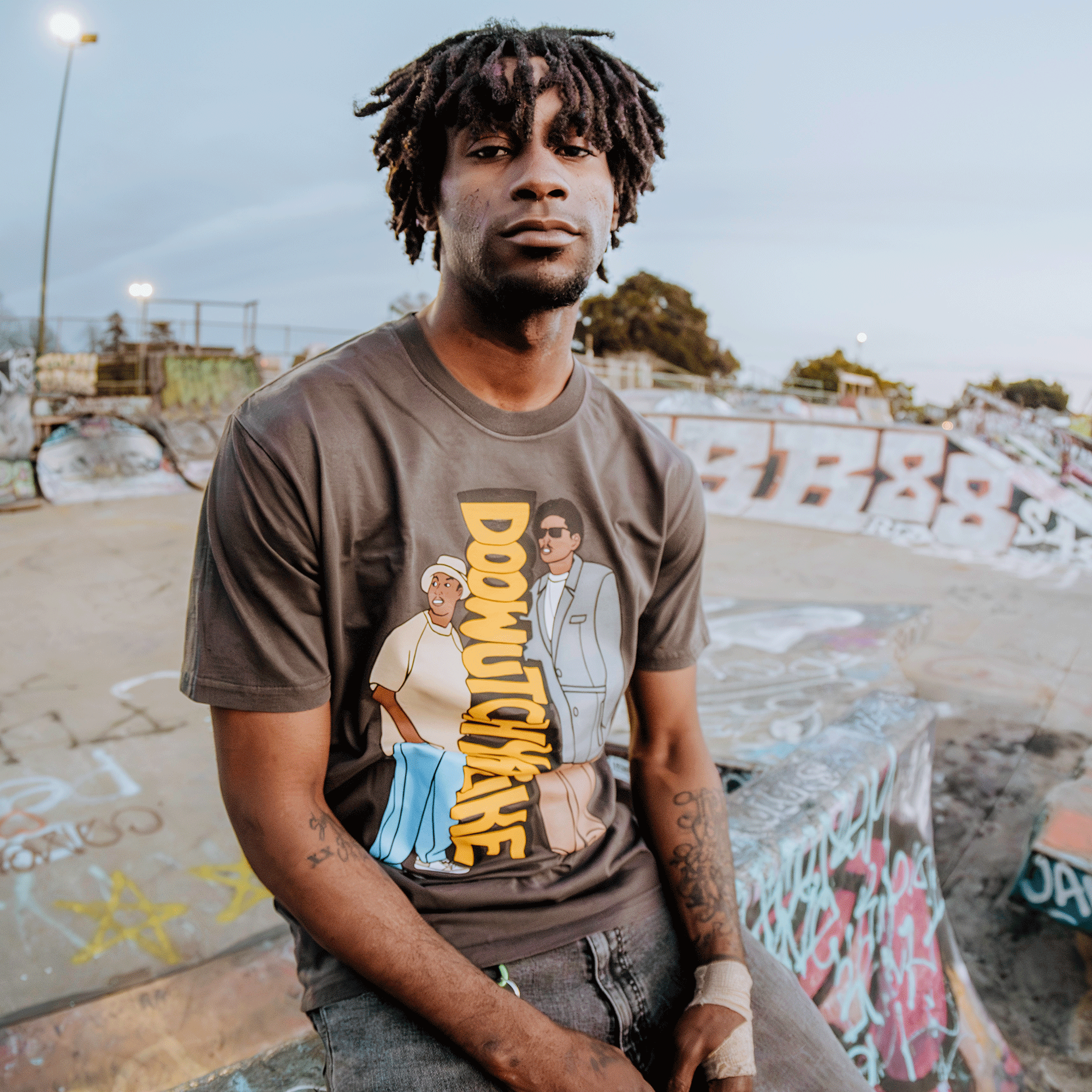 A man in an "Oaklandish Doowutchyalike Tee" with a cartoon design sits in a graffiti-covered skate park, showcasing 90s vibes. With tattooed forearms on display, the scene echoes a Digital Underground aesthetic amidst vibrant graffiti and a clear sky.