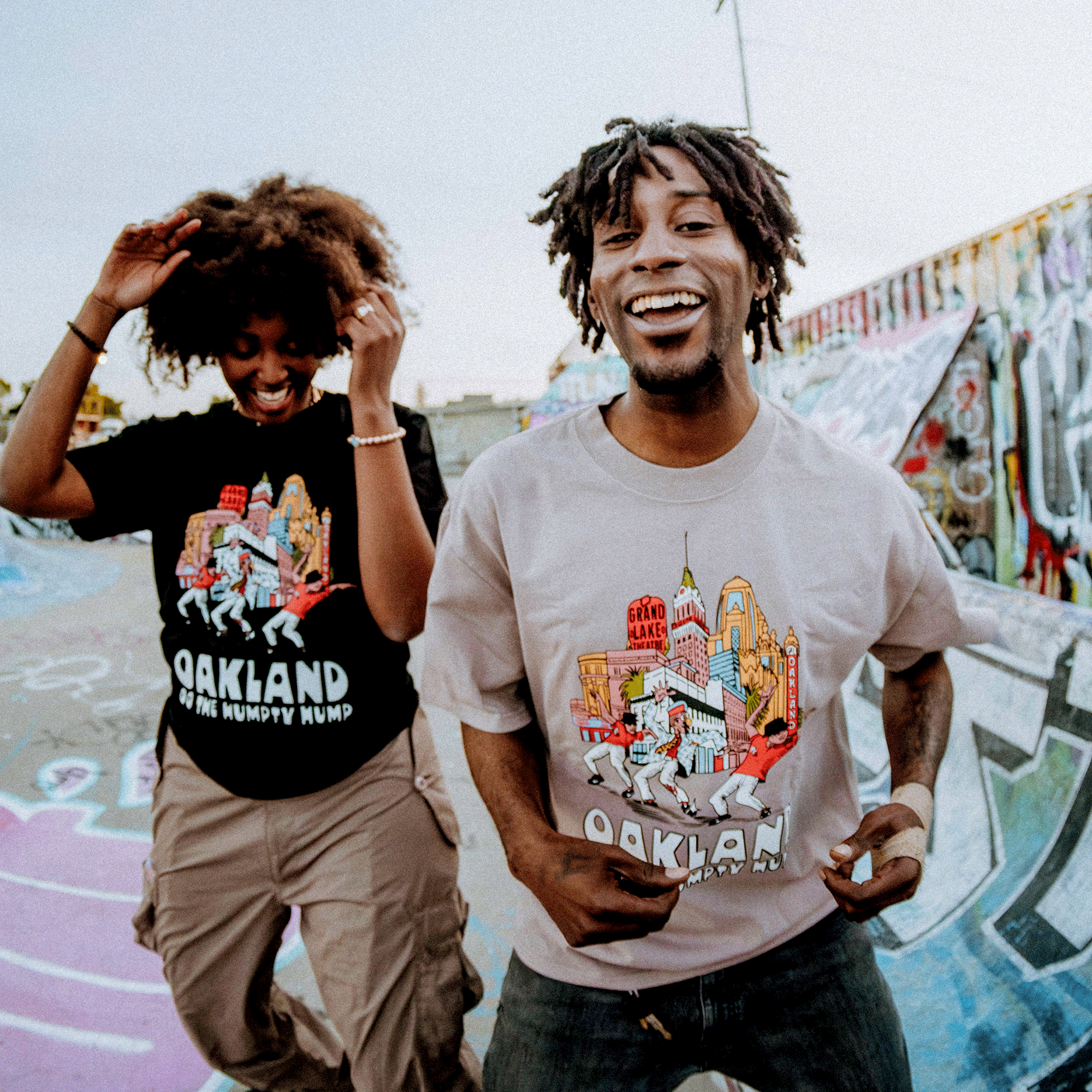 Two people smile and dance on a graffiti-covered rooftop, both wearing Oaklandish "Do the Humpty Hump" tees. The vibrant abstracts behind them pop under the clear sky, while the person on the left, with curly hair and beige cargo pants, channels Humpty Hump vibes.