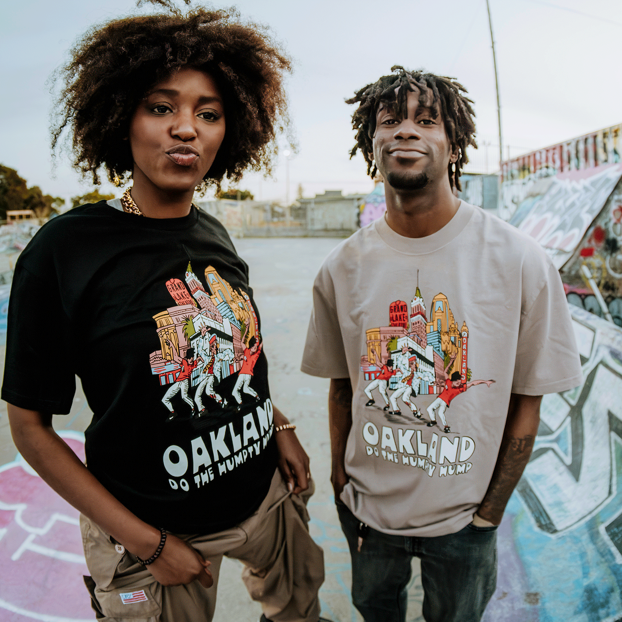 Against a graffiti backdrop, two individuals pose confidently in graphic tees that proclaim "Do the Humpty Hump" by Oaklandish. The one on the left sports an afro, while short dreadlocks adorn the individual on the right, both standing firmly on concrete.