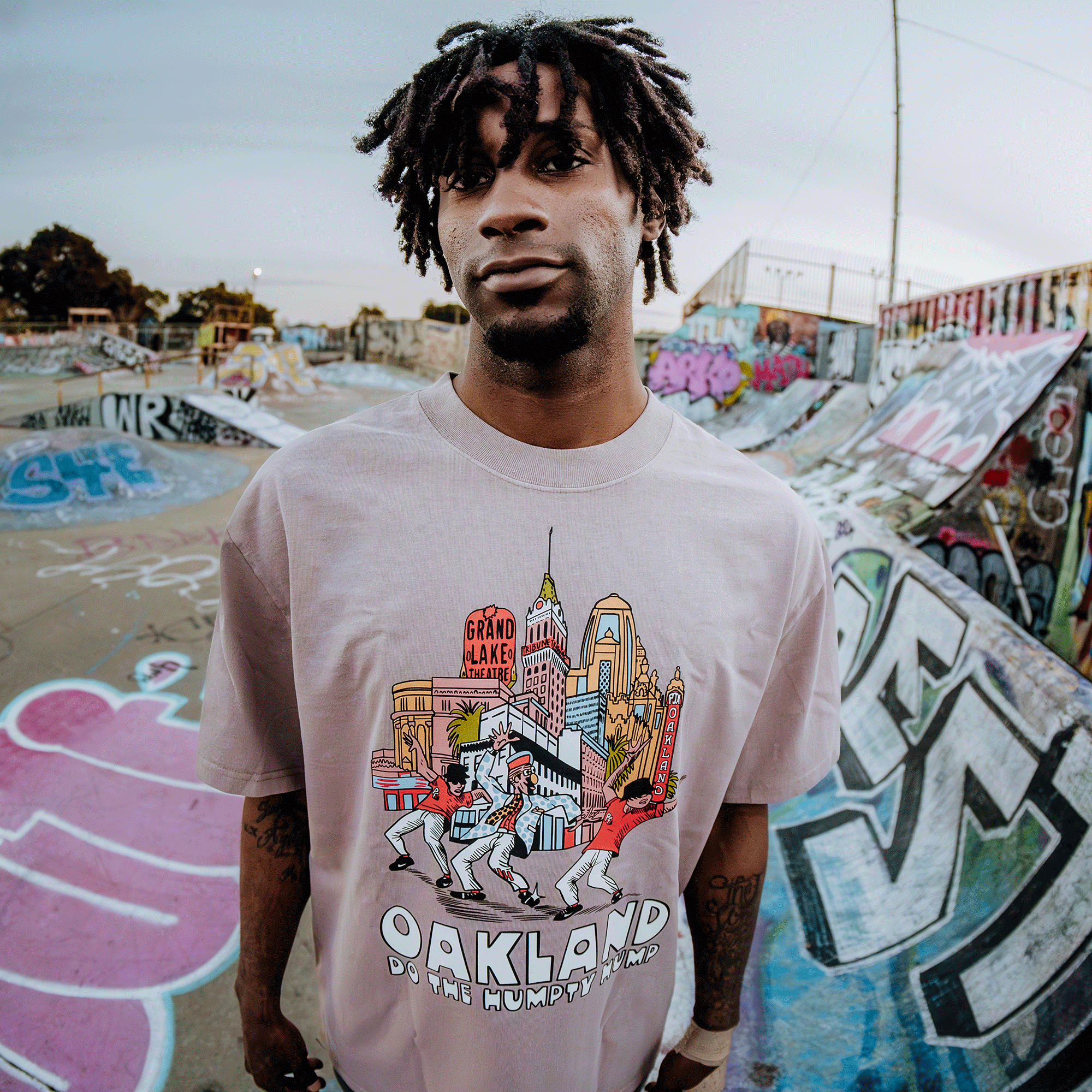 In a graffiti-laden skatepark, a young man wears Oaklandish’s "Do the Humpty Hump" Heavy Tee, featuring a bright cityscape with "Oakland" and "The Humpty Dance," celebrating Digital Underground's Humpty Hump. His short dreadlocks highlight his neutral expression against the vivid urban art.