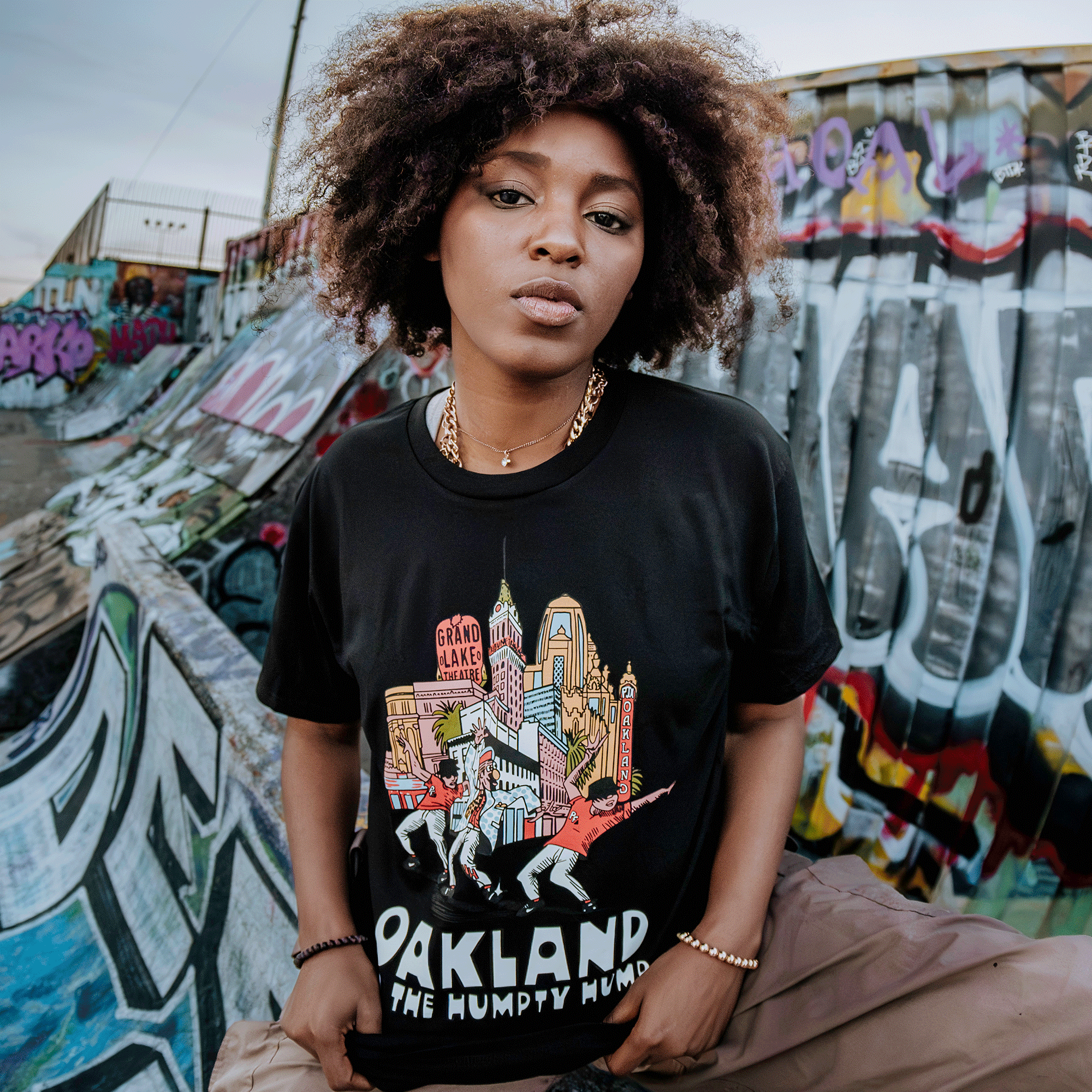 In front of a colorful graffiti wall, a person with curly hair radiates confidence while wearing Oaklandish's "Do the Humpty Hump Tee," a black shirt celebrating Oakland landmarks. With hands in pockets and cool accessories, they embody hip-hop history against an urban sky backdrop.