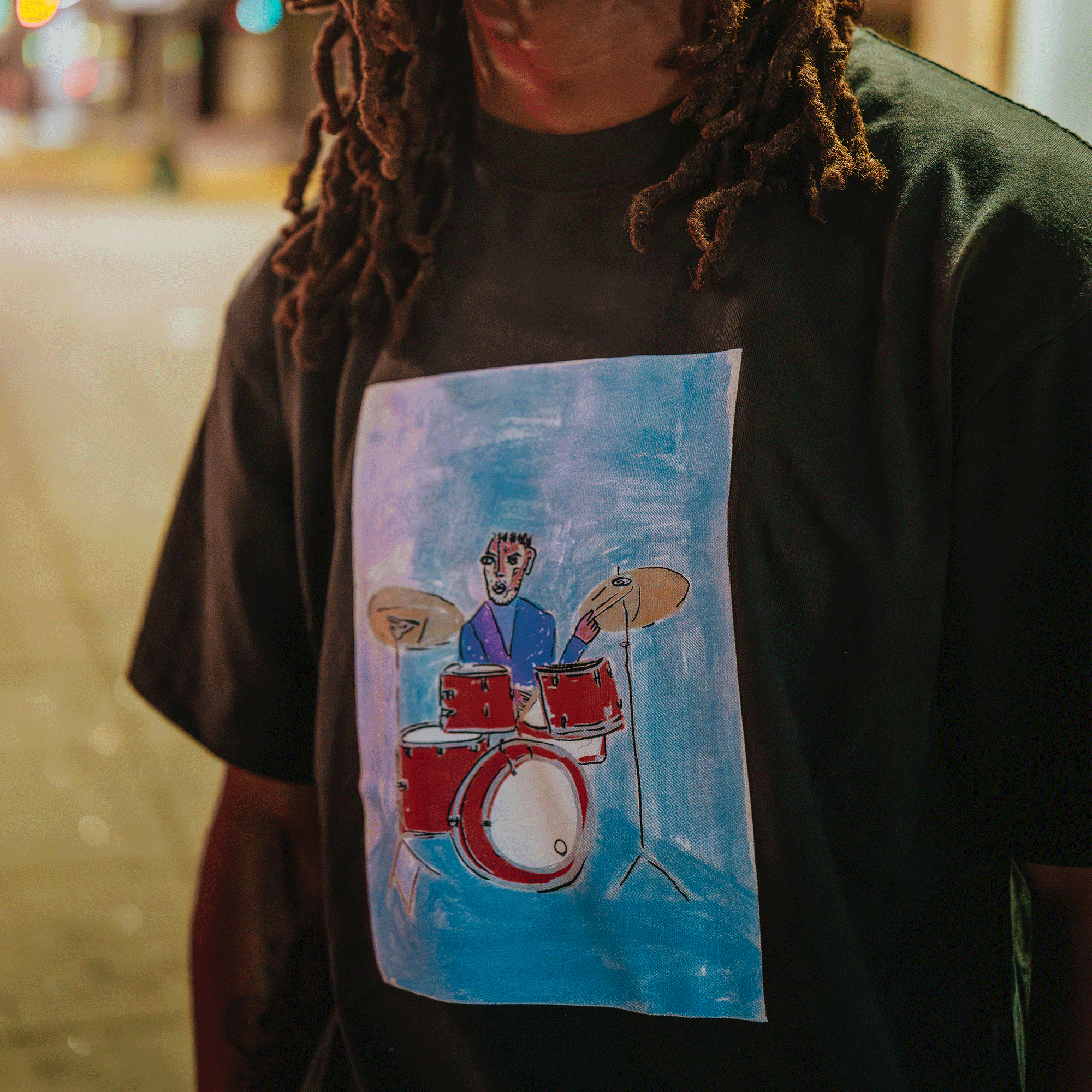 Person wearing a black t-shirt with a colorful drawing of a person playing drums on a blue background.