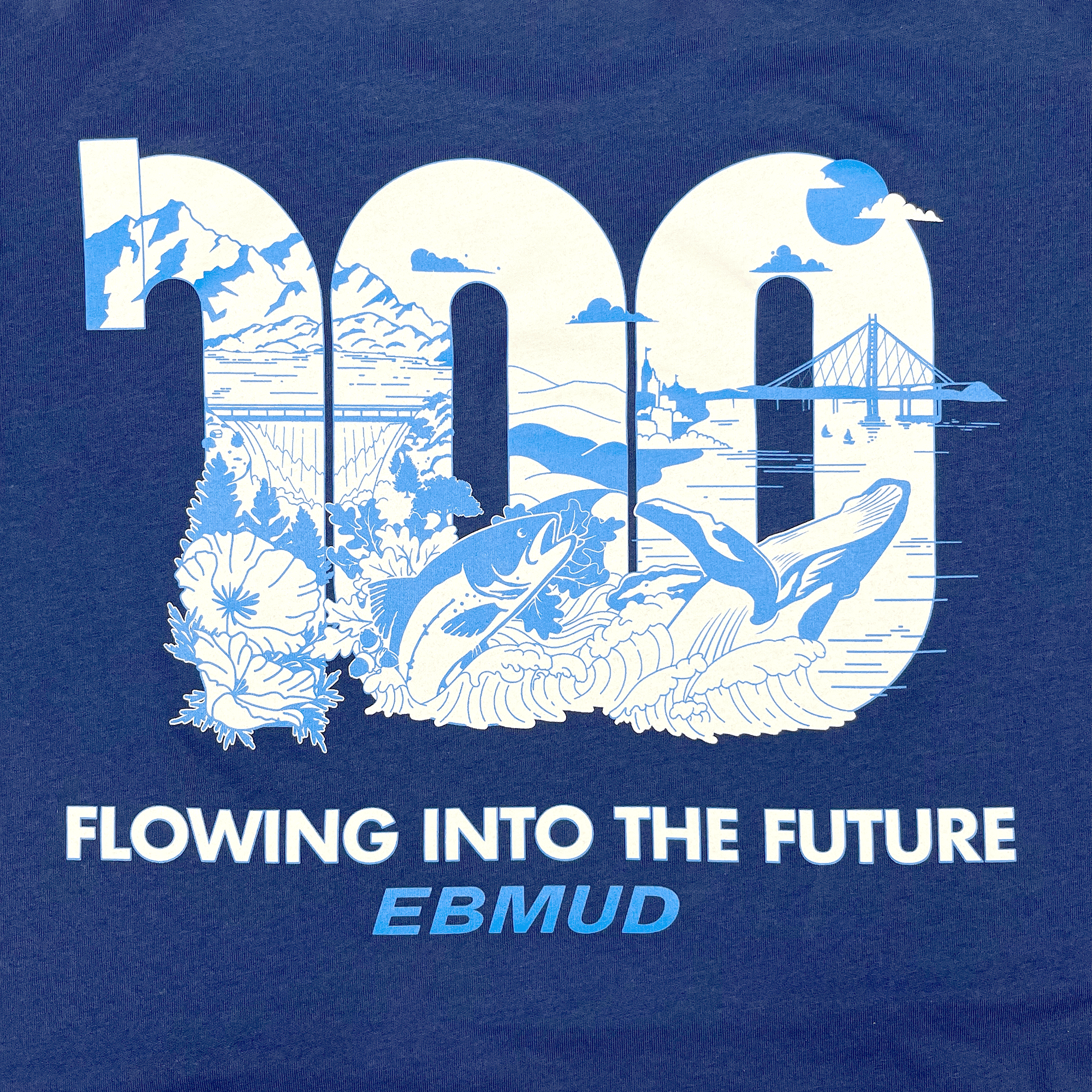 Detailed close-up of EBMUD wordmark and centennial celebration graphic on right chest wear side of blue t-shirt.