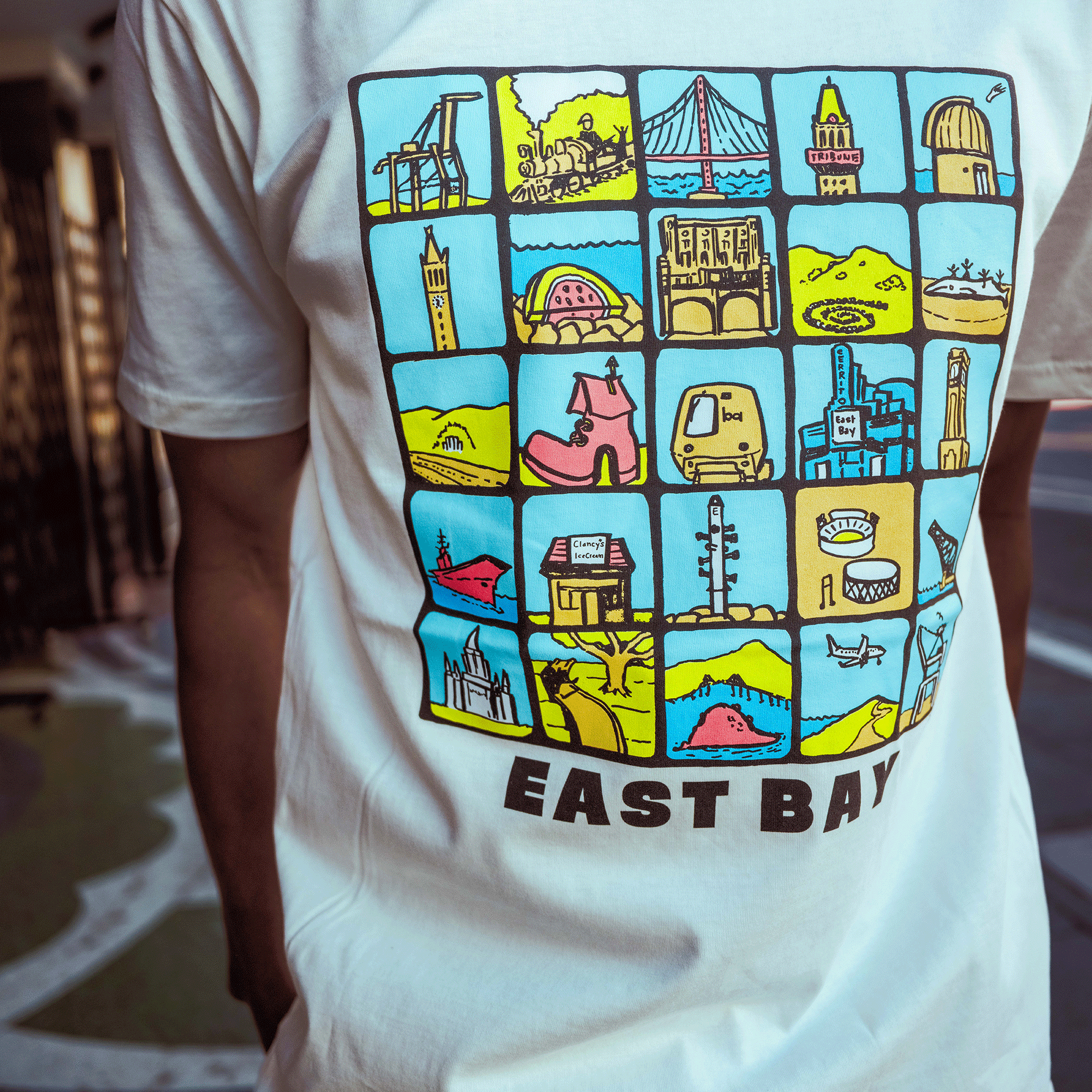 Someone wears the Oaklandish East Bay Landmarks Tee, a white shirt showcasing colorful cartoon squares of Bay Area bridges, buildings, and whimsical scenes on blue and yellow backgrounds.