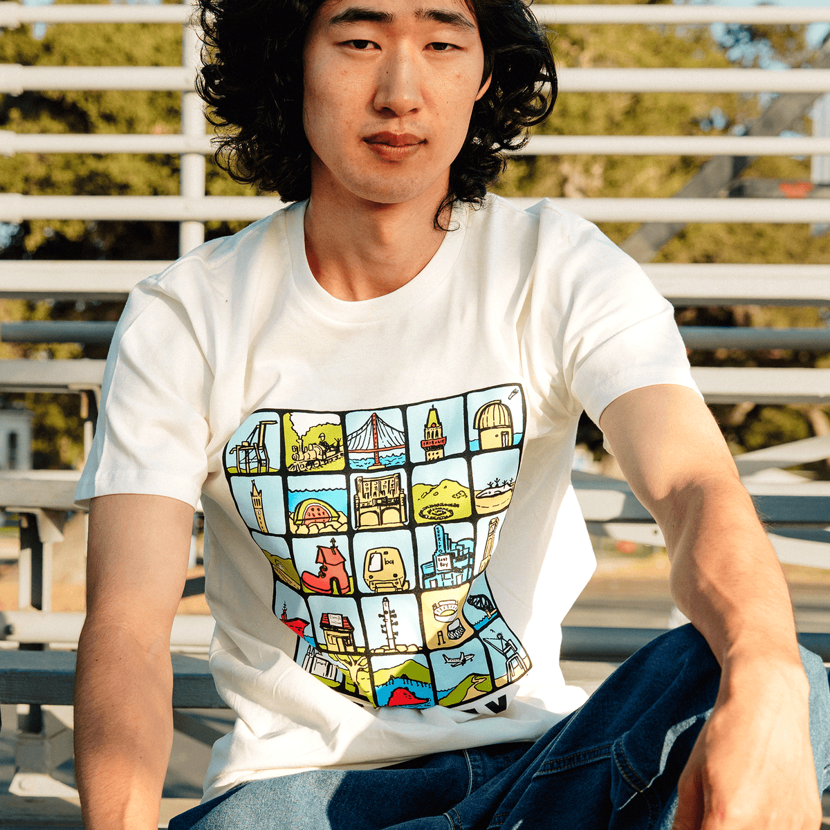 A person with shoulder-length dark wavy hair sits on sunlit metal stadium bleachers wearing blue jeans and an Oaklandish East Bay Landmarks Tee, a cream-colored t-shirt featuring an East Bay grid of colorful botanical art landmarks.