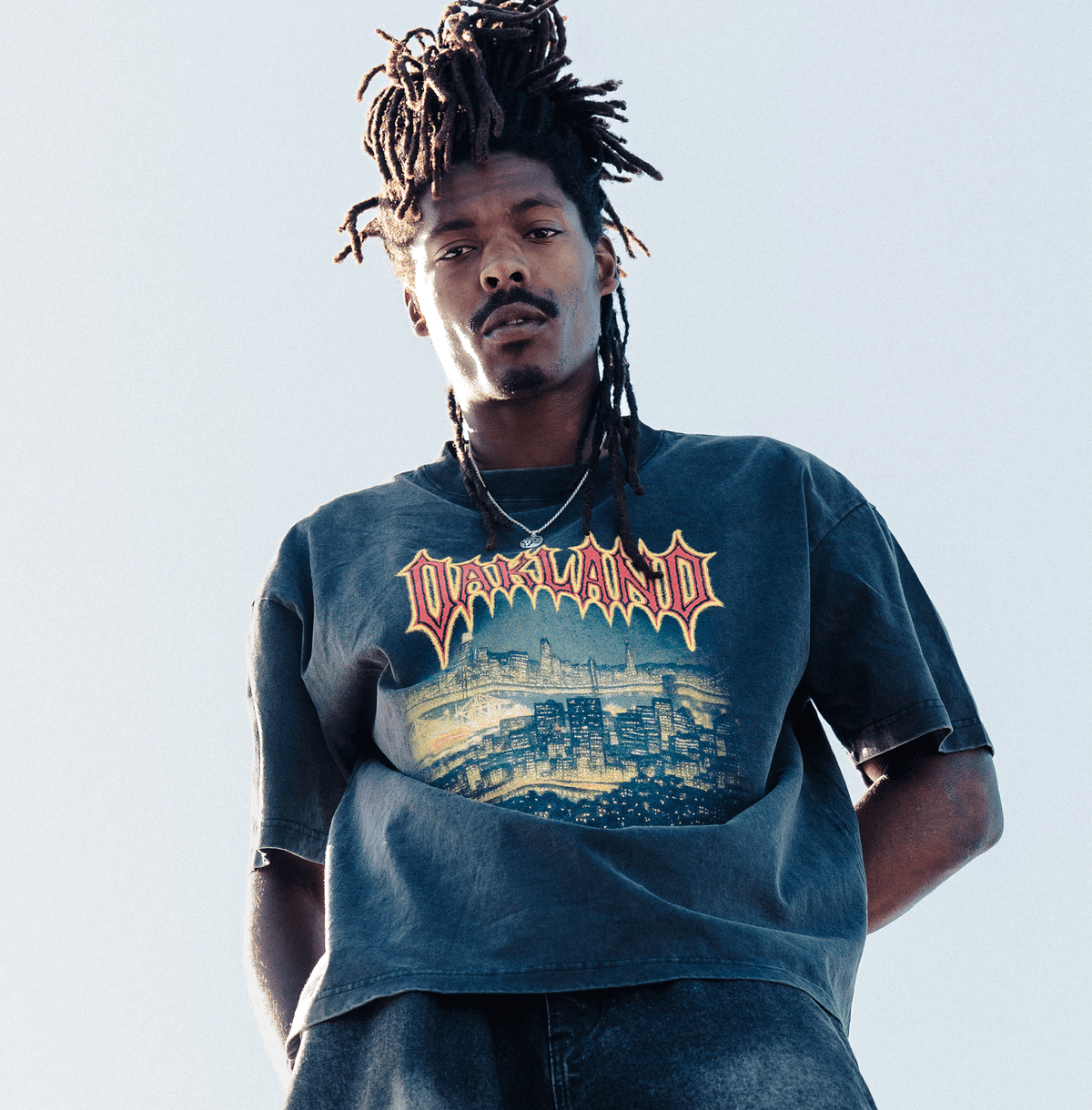 A person with dark locs and a mustache wears an Oaklandish Ember Tee featuring a yellow and red city skyline graphic with “OAKLAND” in metal font, shot from a low angle against a clear sky.