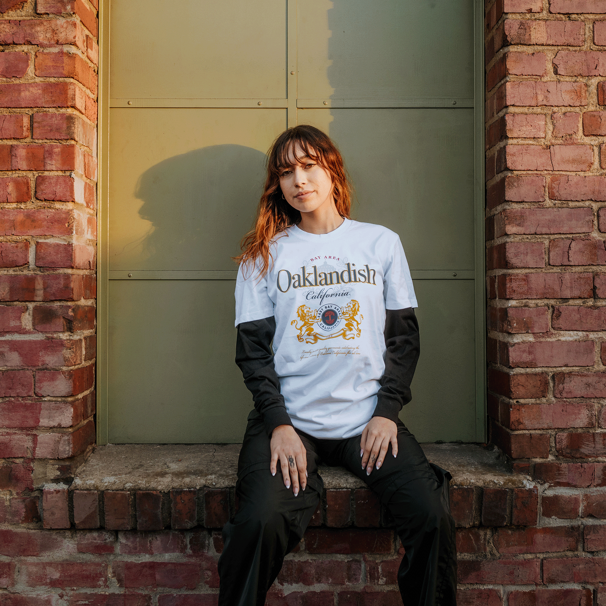 A young woman with wavy hair sits on a brick windowsill, wearing the Oaklandish Lager Especial Tee over a long-sleeve black shirt and black pants. The red brick wall and closed metal window create a calm backdrop as she gazes at the camera.