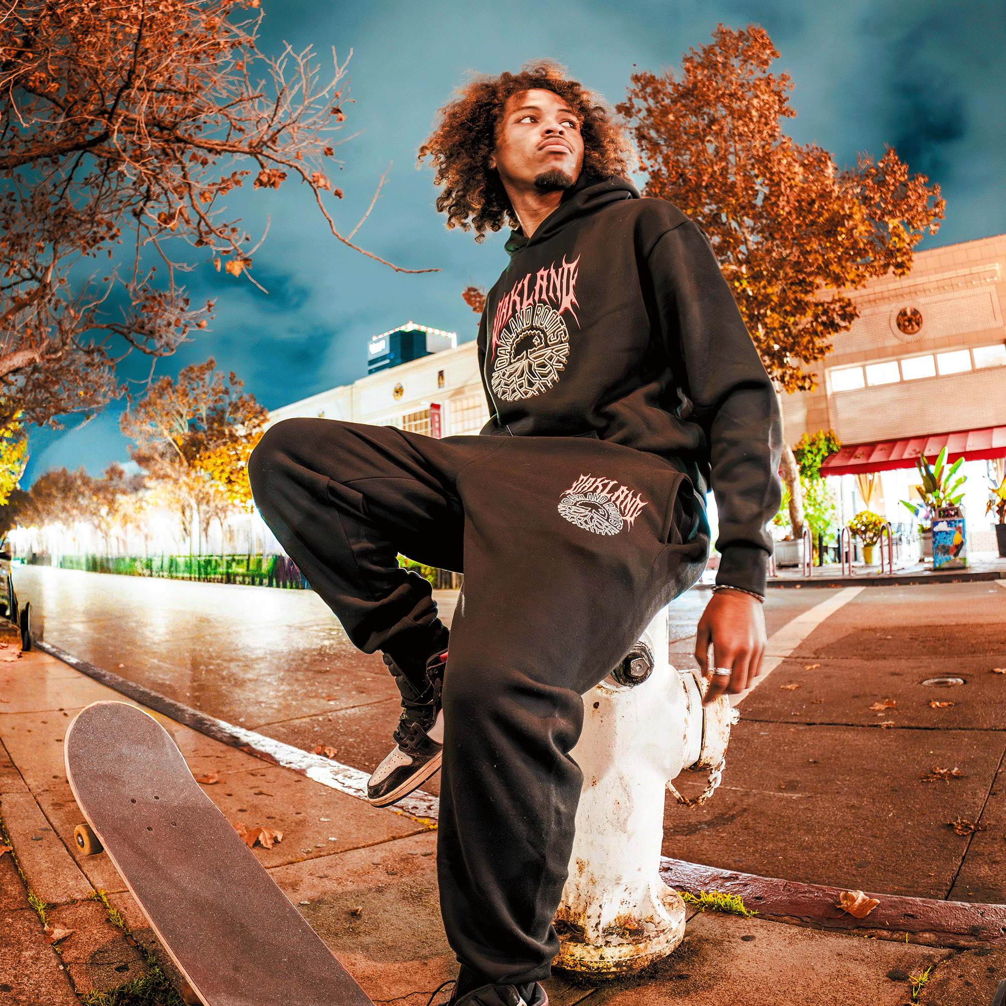 A young man with curly hair wears an Oakland Roots SC Forge Hoodie and sweatpants, sitting on a fire hydrant at night with one foot on a skateboard. He looks to the side as autumn trees and an empty street glow behind him.