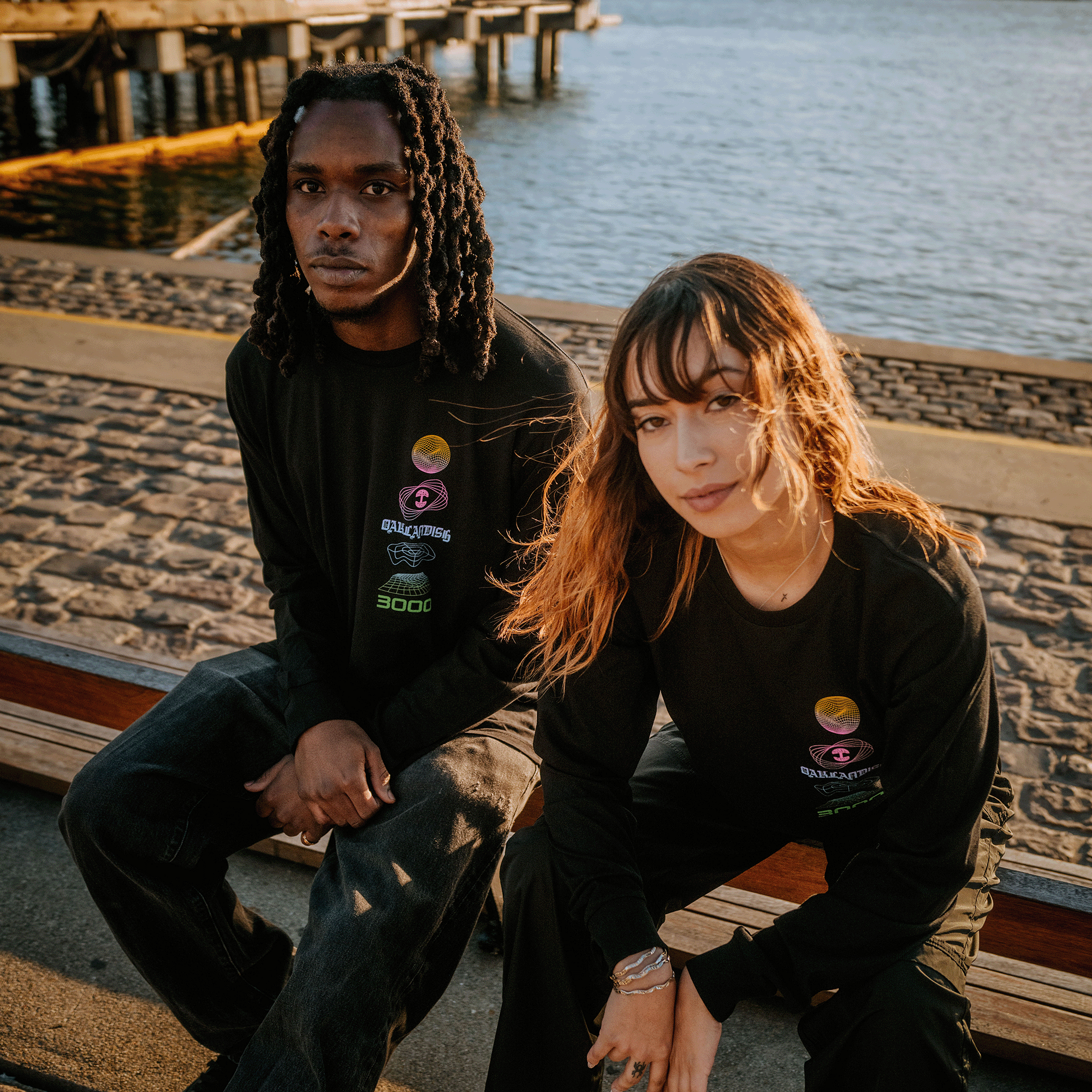 Female and male model wearing long-sleeve t-shirt with neon pink, yellow, blue, and green futuristic graphic with OAKLANDISH wordmark.