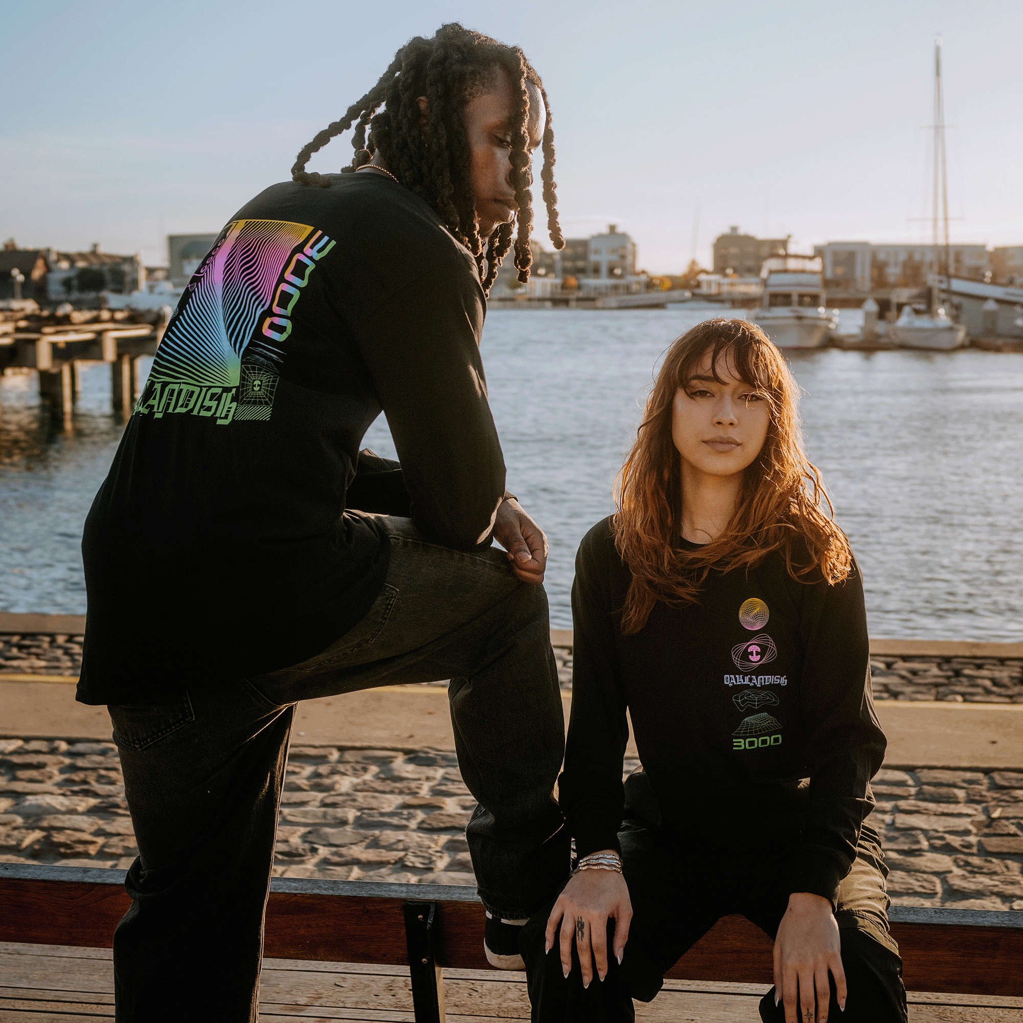 Female and male model wearing long-sleeve t-shirt with neon pink, yellow, blue, and green futuristic graphic with OAKLANDISH wordmark.