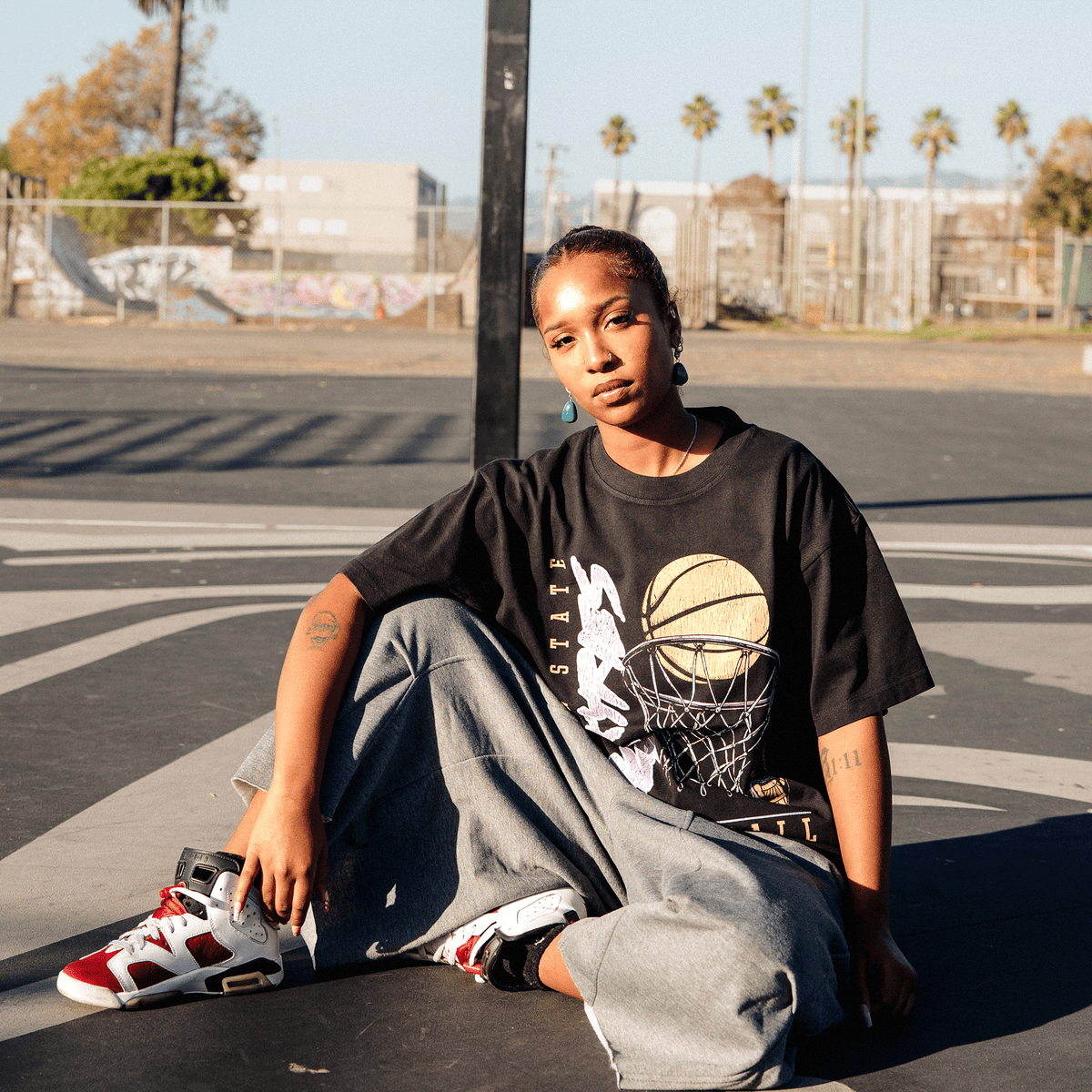 A person with their hair pulled back sits on an outdoor basketball court wearing a classic fit GSV Swish Tee, loose gray trousers, and red and white sneakers. They look directly at the camera with a neutral expression, with palm trees visible in the background.