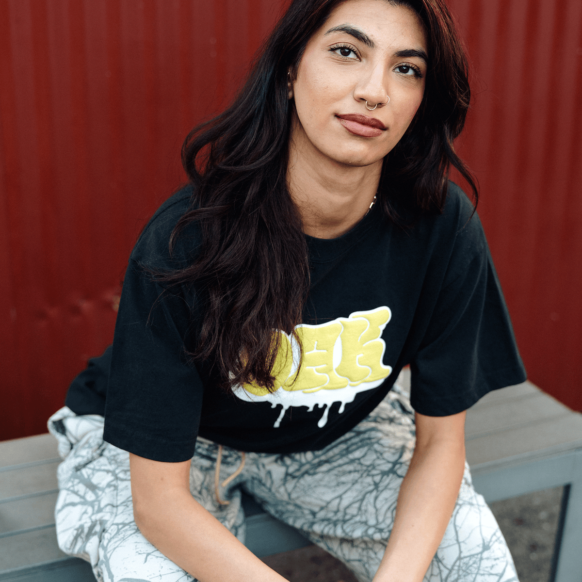A person with long, dark wavy hair and a septum piercing wears an Oaklandish Graff OAK Tee with a yellow graffiti-style graphic while sitting on a light-colored bench in front of a deep red corrugated metal wall, wearing patterned grey and white pants.
