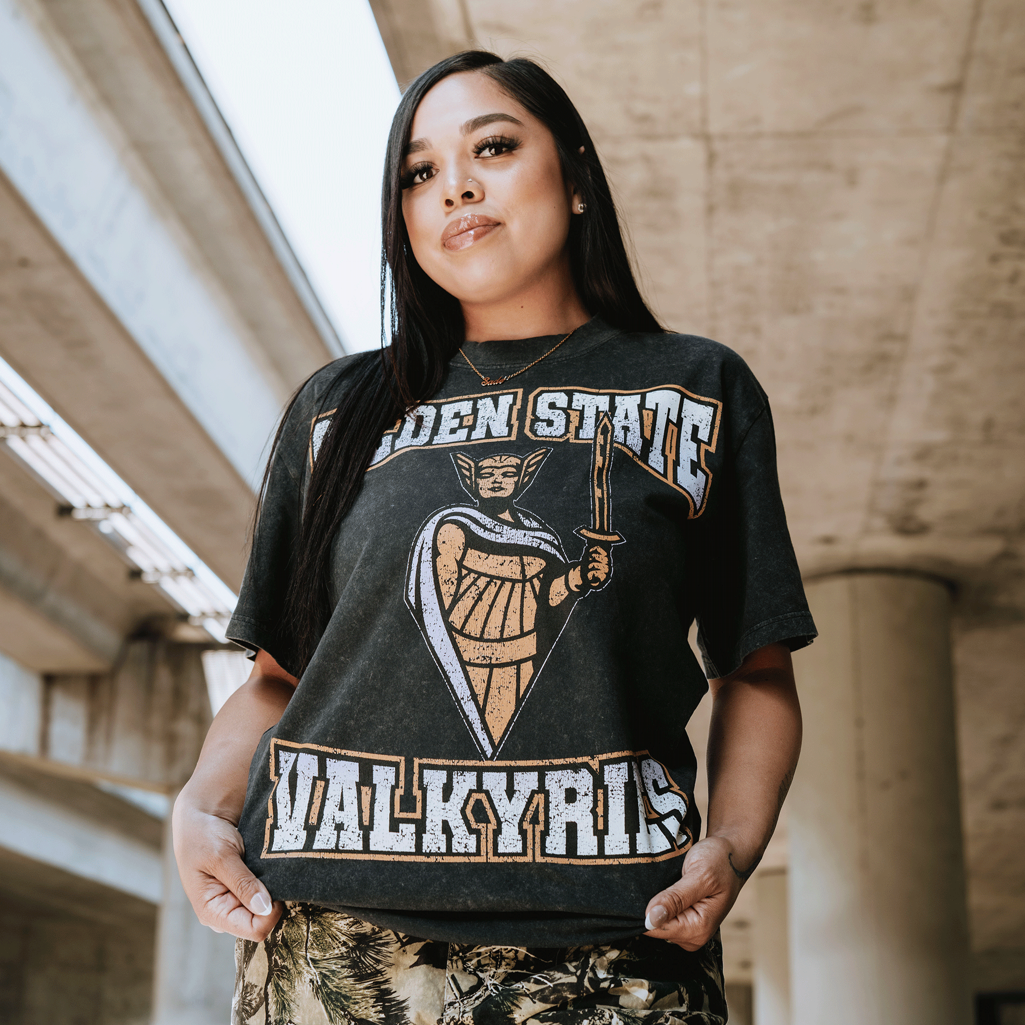 A woman with long dark hair stands confidently under an overpass, wearing the GSV Golden State Valkyries Guardian Stone Wash Heavy Tee with a warrior graphic and patterned pants. Sunlight highlights her relaxed pose and neutral expression.
