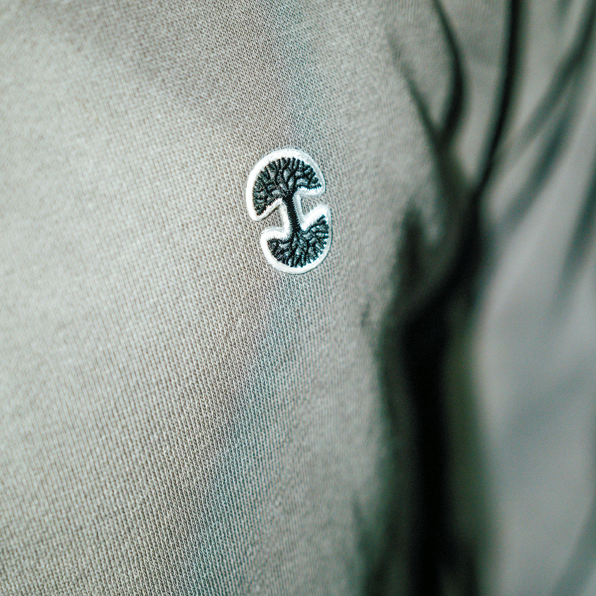 Close-up of the Oaklandish Hojicha 1/4 Zip's grey, garment-dyed fabric, showcasing a small off-center embroidered logo—a stylized tree in dark green and white within a circle—on mid-weight outerwear.