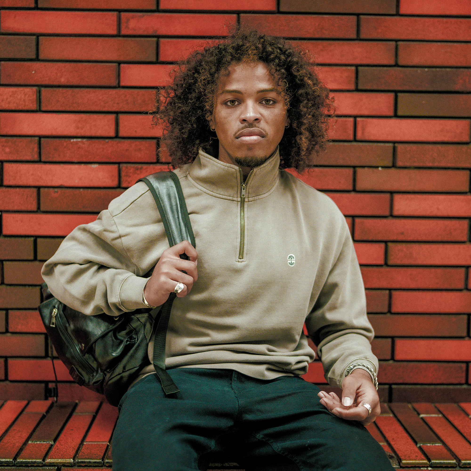 A young man with curly hair sits on a bench by a red brick wall, wearing the Oaklandish Hojicha 1/4 Zip pullover and dark pants. He carries a black backpack over one shoulder, looking directly at the camera with a neutral expression.