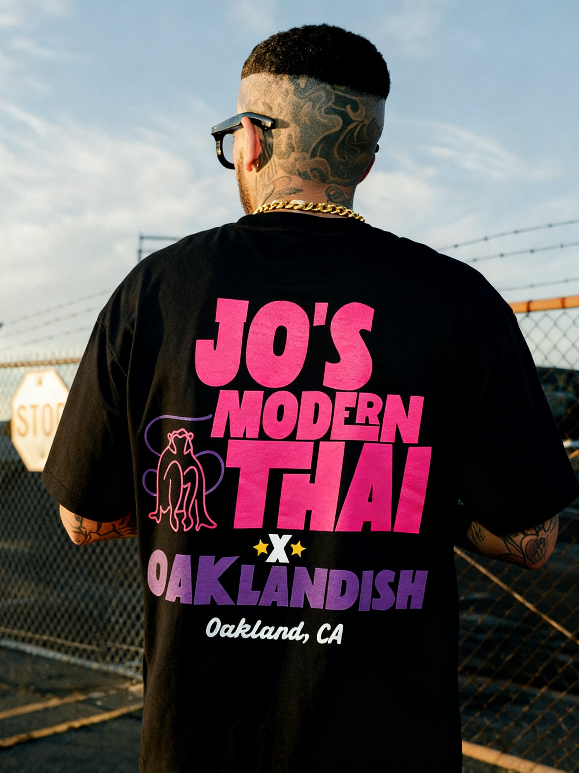 A tattooed person in sunglasses faces away, wearing the Jo's Modern Thai X Oaklandish Tee by Oaklandish—a heavy-weight black T-shirt with bold pink and purple text, plus two elephants and a fence graphic in the background.