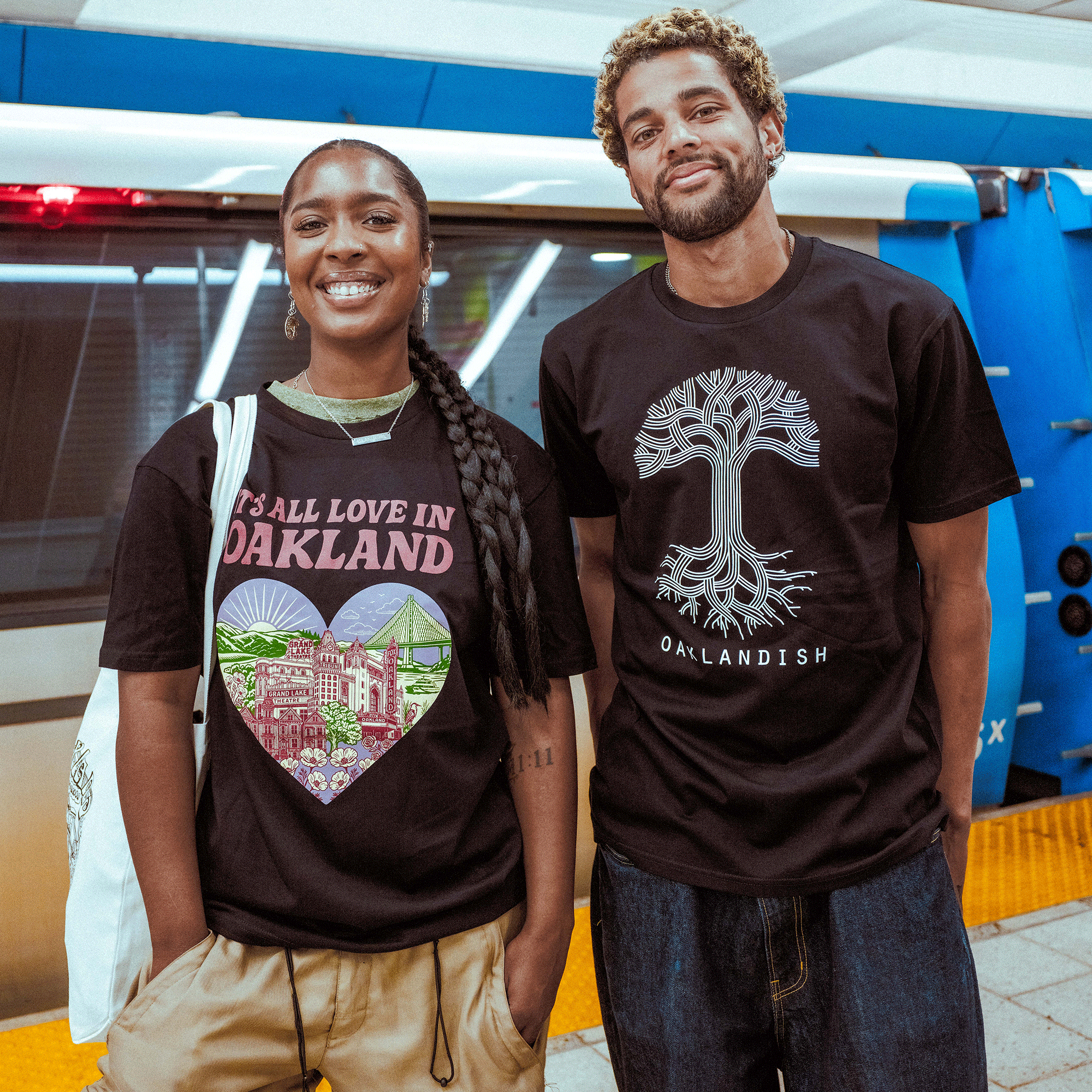 Two people smile on a subway platform. She wears the black "It's All Love in Oakland Tee" by Oaklandish, featuring colorful landmarks. He sports a black Oaklandish shirt with a white tree graphic. A blue train is stopped behind them.