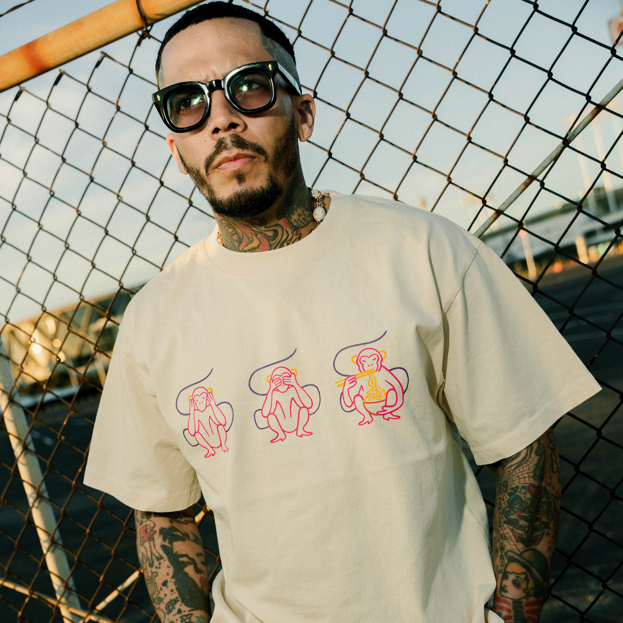 A tattooed man wearing sunglasses and the Jo's Modern Thai X Oaklandish Tee by Oaklandish, featuring three colorful cartoon monkeys, stands outdoors in sunlight by a metal chain-link fence, looking away from the camera.