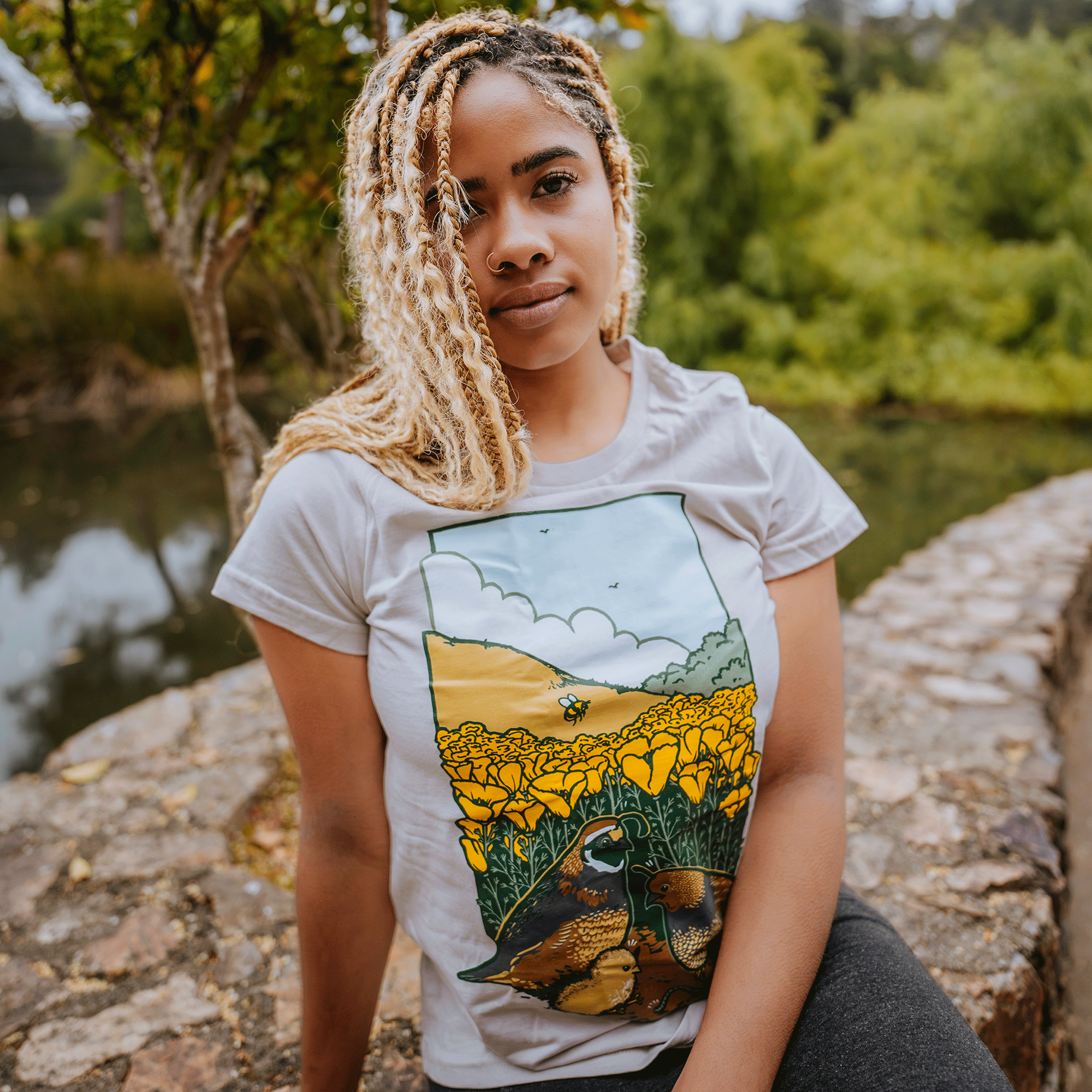 A person with long, braided hair sits on a stone wall near a pond and trees, wearing the Women's Quail Family Tee by Other. The 100% cotton classic fit t-shirt is adorned with yellow flowers and ducks, enhancing the serene scene filled with lush greenery in the background.