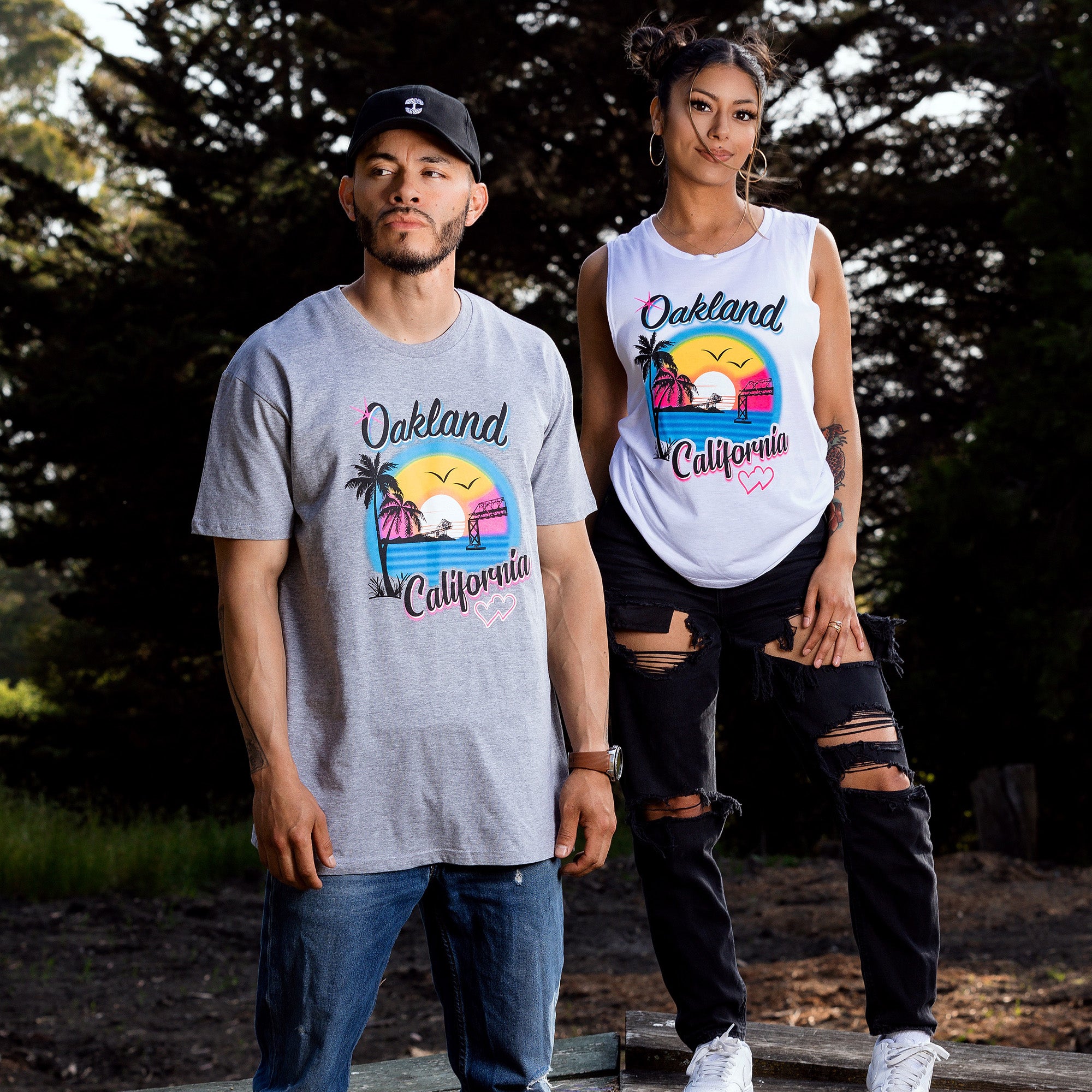 Male and female model standing outdoors wearing grey tee and white tank top with multicolor bright ink colors coastal bay scene with palm trees and bridges and hearts, text spelling Oakland, California.