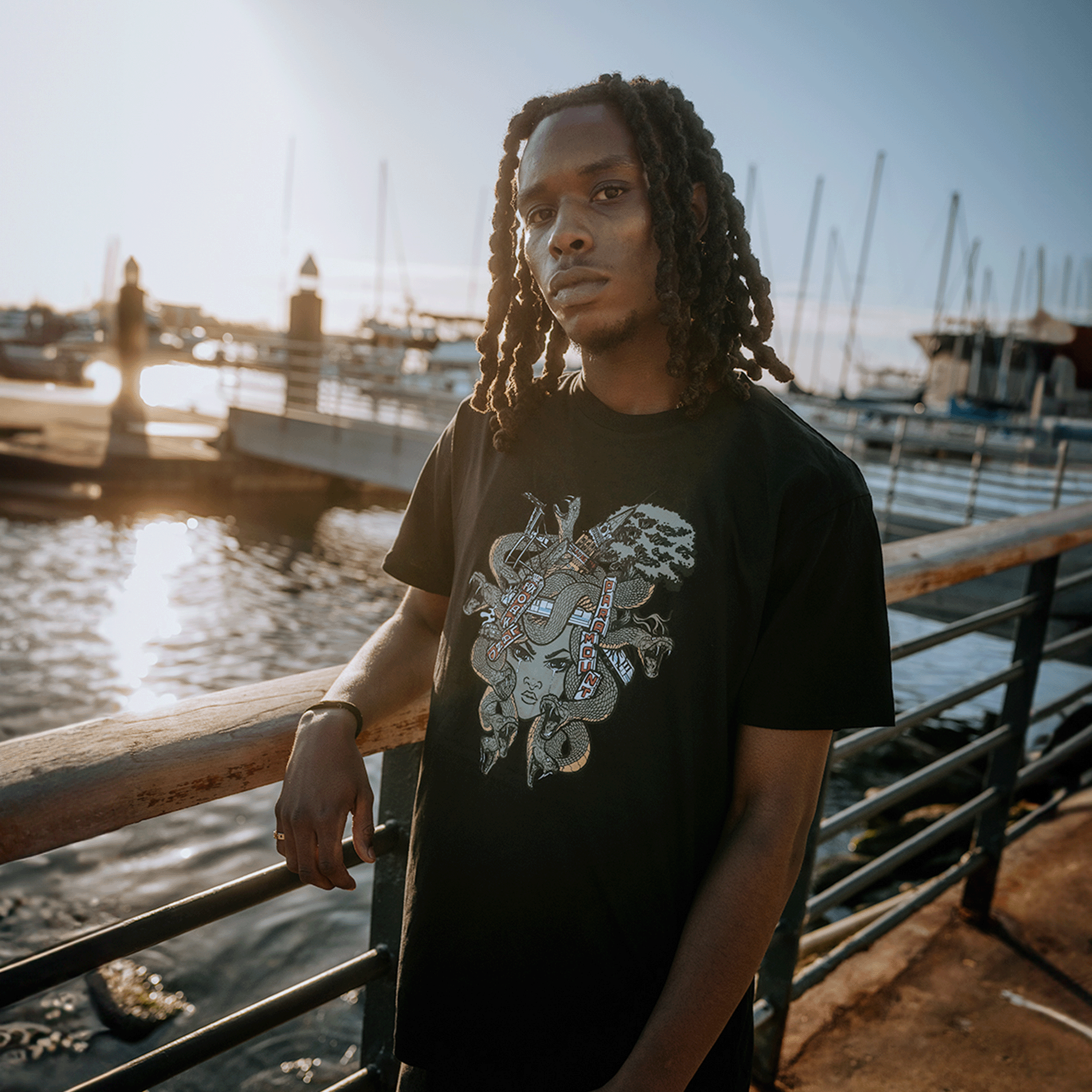 A person with long, braided hair stands against a metal railing by the water, with boats and sailboats in the background. They are wearing an Oaklandish Medusa Tee from the men's collection, made of 100% cotton and featuring a colorful graffiti-style design. The sunlight creates a soft glow, enhancing the serene waterside atmosphere.