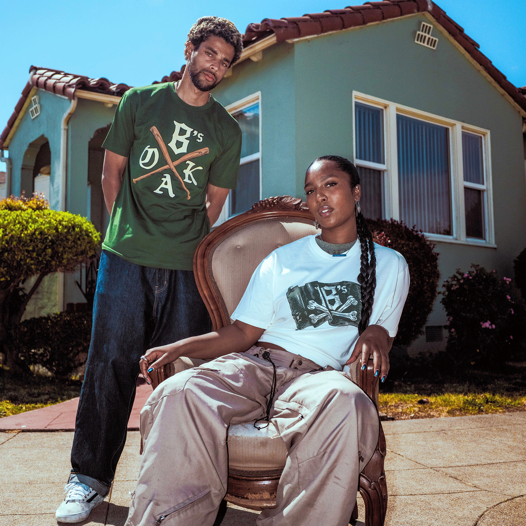 A man stands behind a woman seated outdoors by a light green house. He wears the Oakland Ballers Jolly Roger Tee with baggy jeans; she has on a white Pioneer League tee and beige pants. Both look relaxed and confident as they pose.