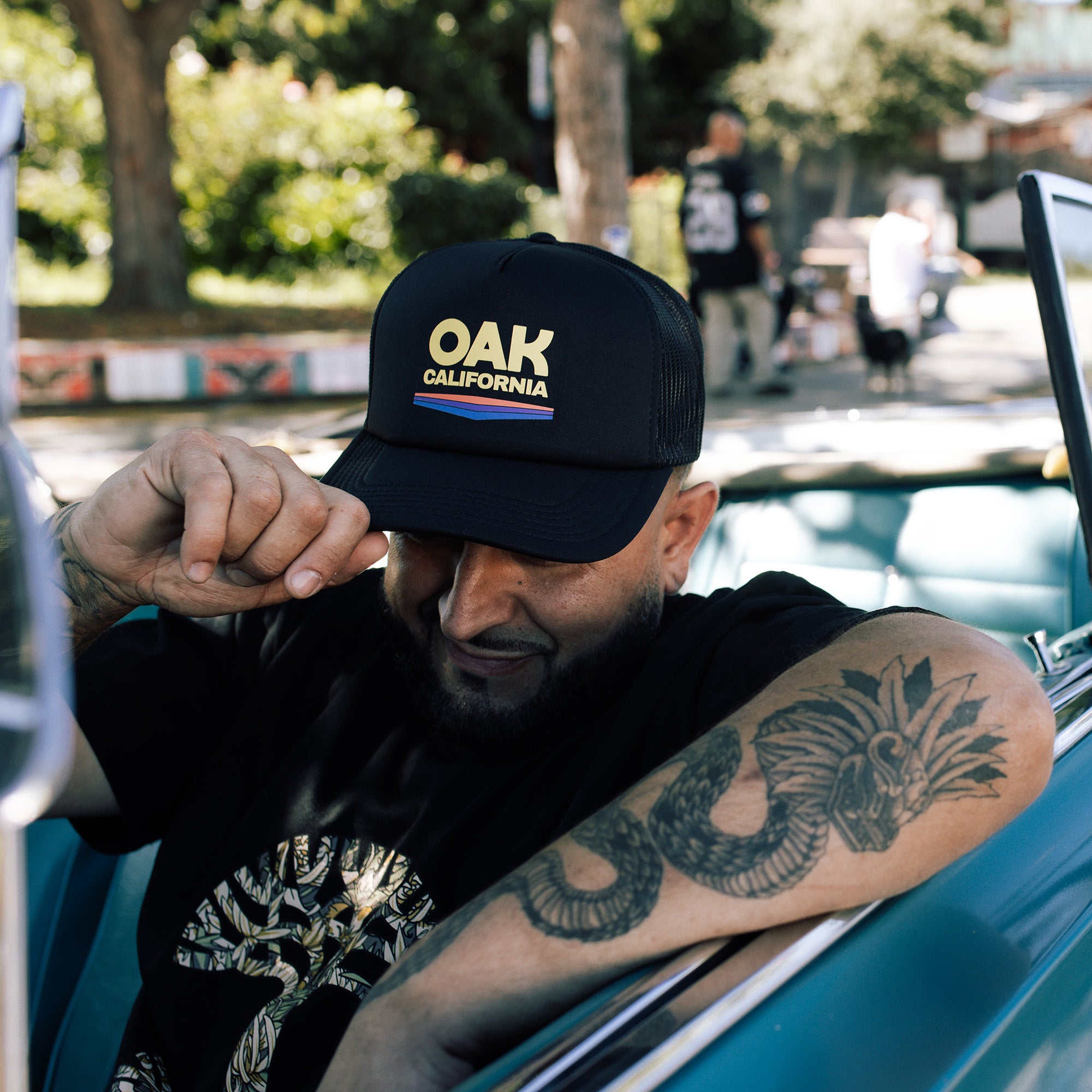 Model wearing black trucker cap with color Oak California graphic on front foam panel with curved brim.