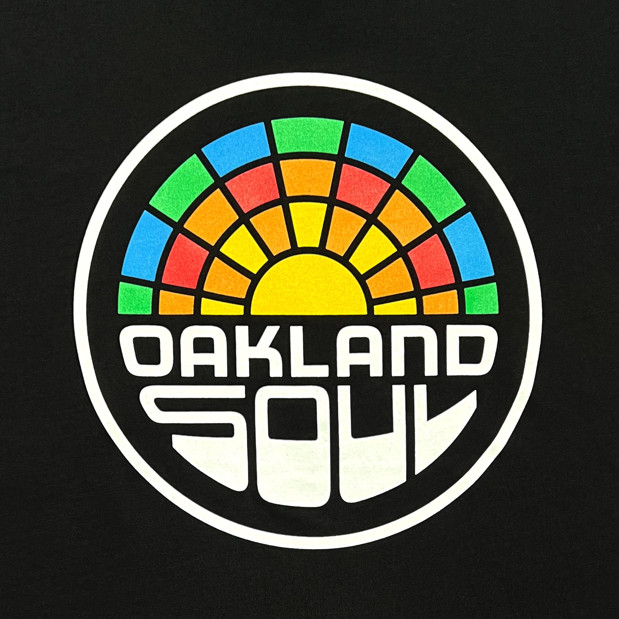 The Oakland Soul Logo Minus Tee by Oakland Soul features a circular logo with a stylized sun at the bottom, blue, green, yellow, and orange segments radiating outward, and bold white "OAKLAND SOUL" text on a black background.