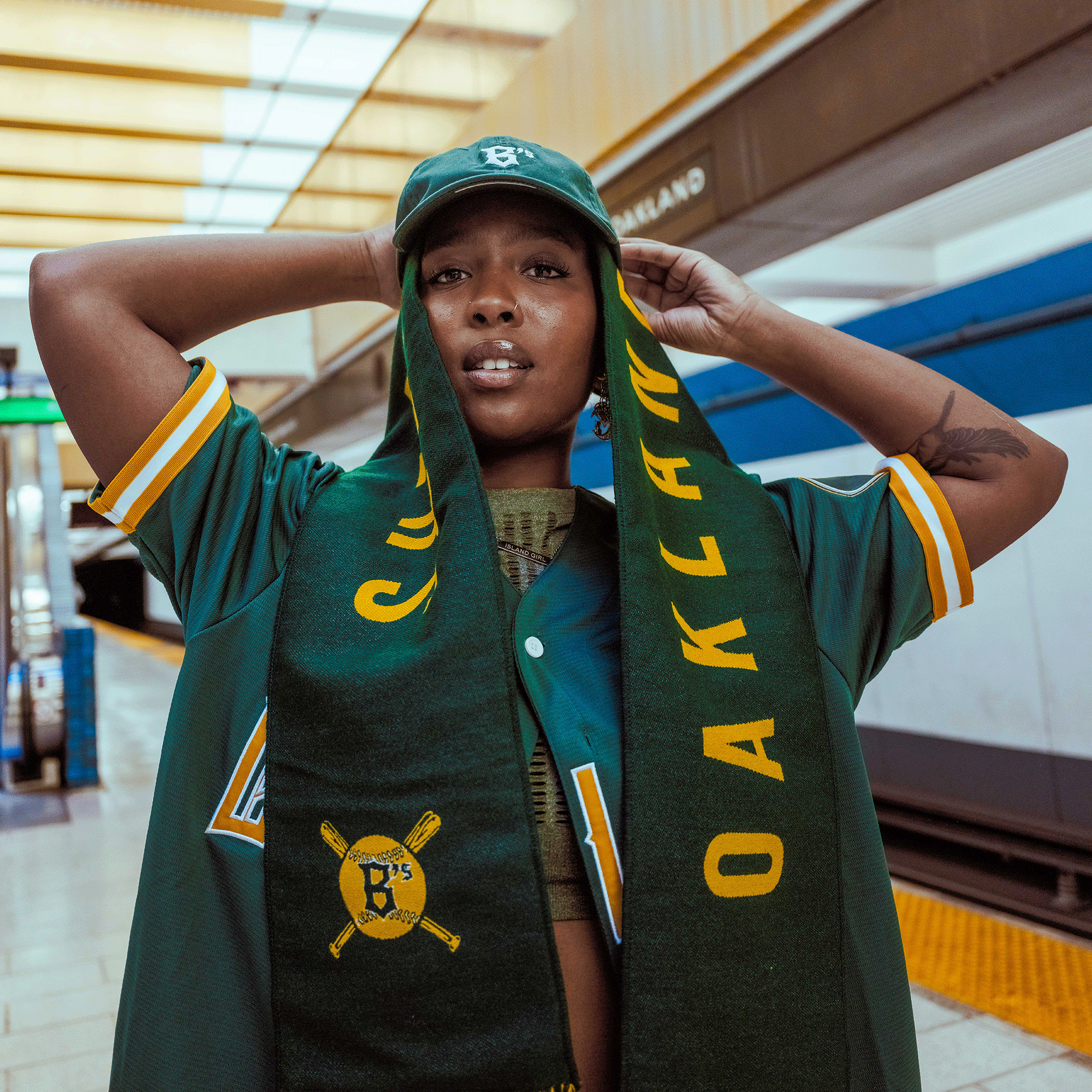 A woman on a subway platform confidently holds up an Oakland Ballers Scarf (by Oakland Ballers) in green and yellow. She wears matching team gear, with a train behind her and "OAKLAND" visible on the station sign.