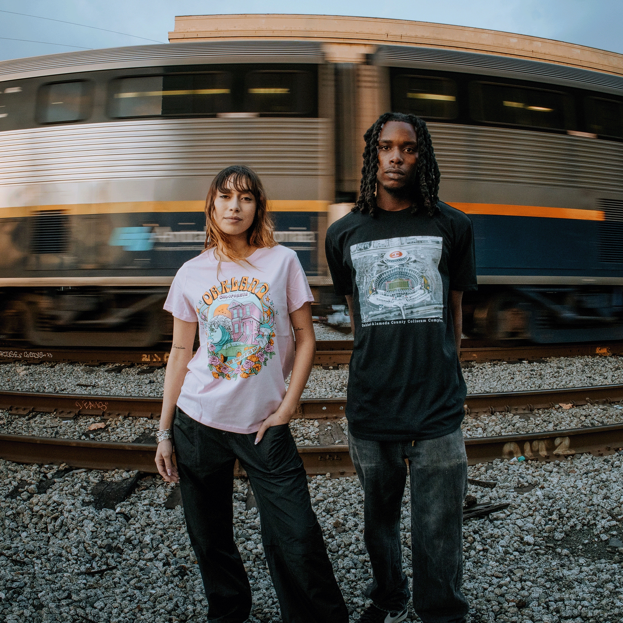 A man and woman are standing outside before a moving train wearing Oaklandish t-shirts.