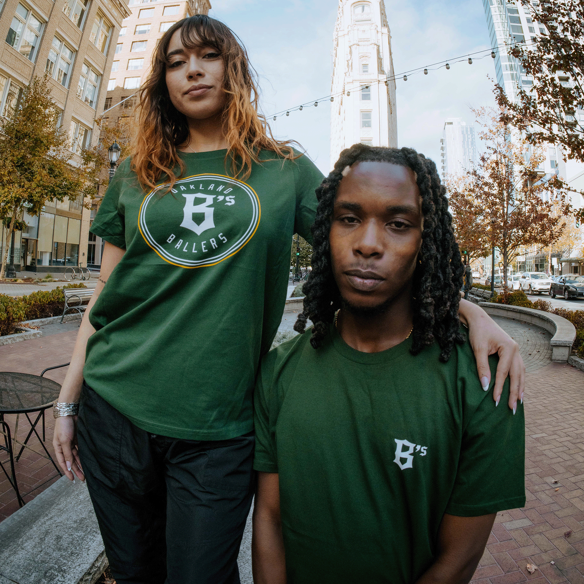 Two individuals stand outdoors in an urban setting with tall buildings and string lights in the background, both wearing green Oakland Ballers t-shirts. The woman on the left, dressed in a Women's Oakland Ballers Logo Tee, has her arm around the shoulder of the man on the right. Both shirts proudly display a logo celebrating their Pioneer League team.