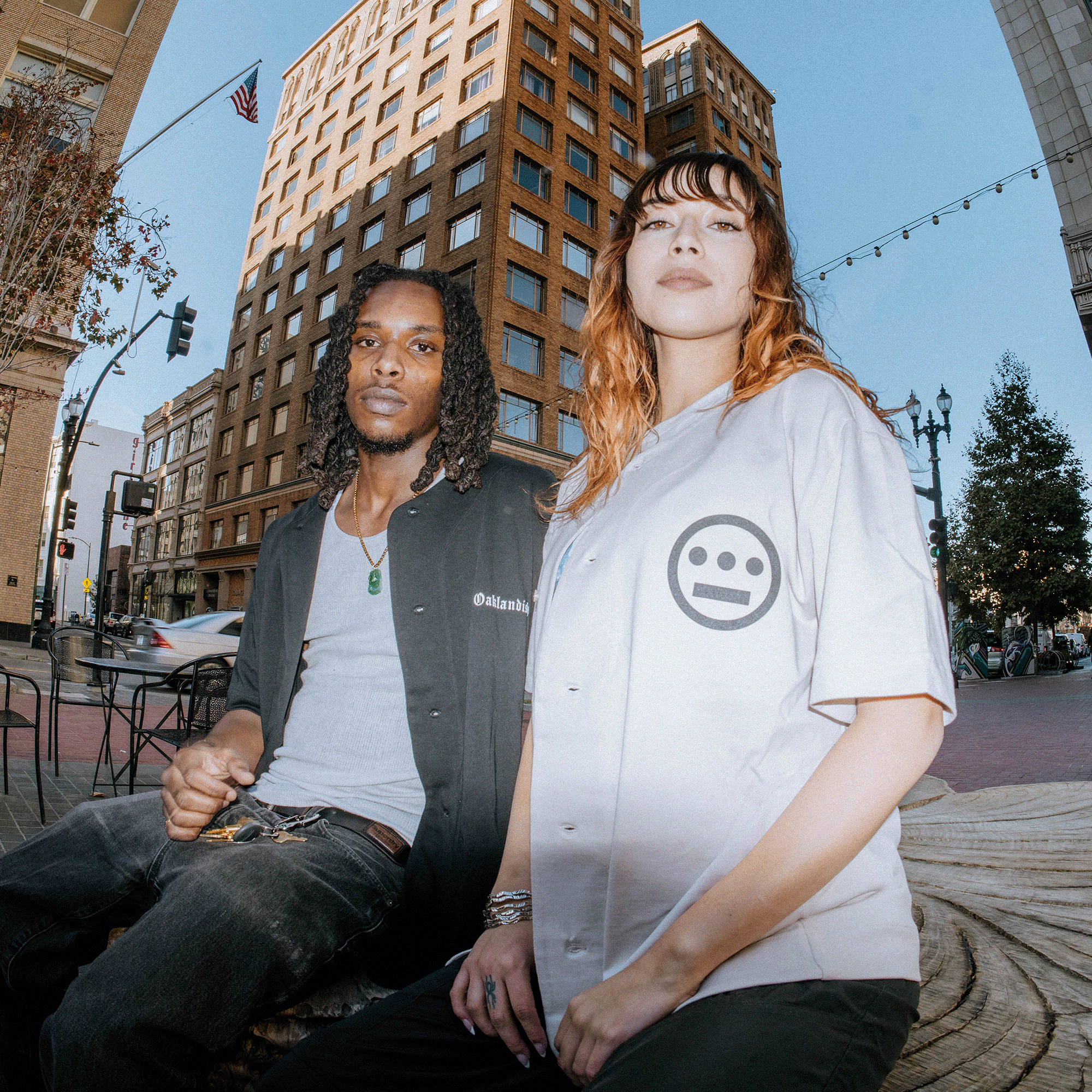 Two individuals sit on a large wooden seat in an urban area, with a tall brick building in the background. The person on the left wears a dark shirt and jeans, while the person on the right is sporting an Oaklandish Heavy Button-up Baseball Jersey from Oaklandish, featuring their iconic tree logo. String lights and trees are partially visible.