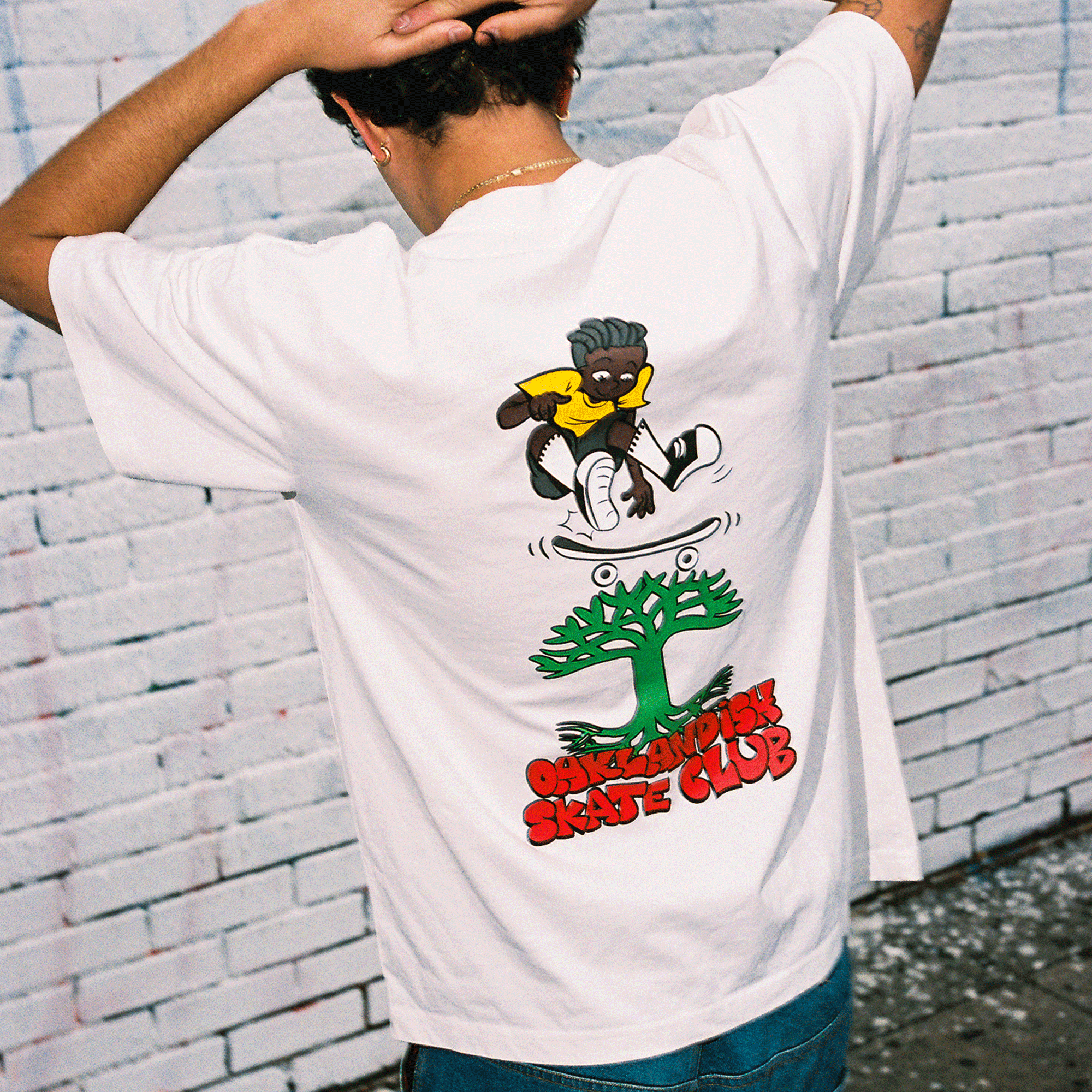 A person stands with their back to the camera, hands on head, wearing the Oaklandish Skate Club Tee—a classic fit by Oaklandish with a cartoon skater above a green tree and bold red "OAKLANDISH SKATE CLUB." The background is a white brick wall.