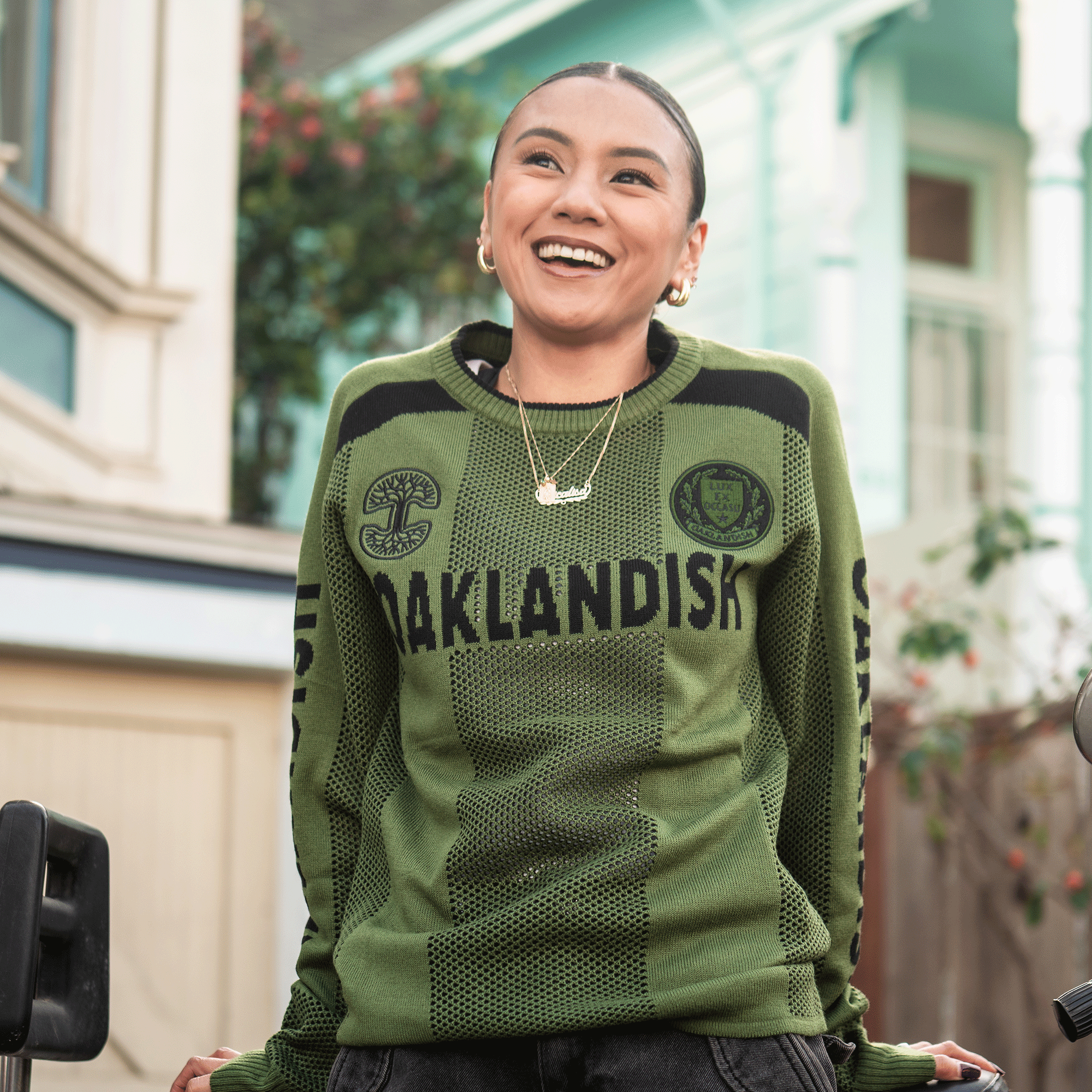 A smiling person stands outdoors in front of pastel houses, enjoying the sun in a green Oaklandish Terrace Sweater with black text and designs, featuring "OAKLANDISH" across the chest, styled in men's sizing.