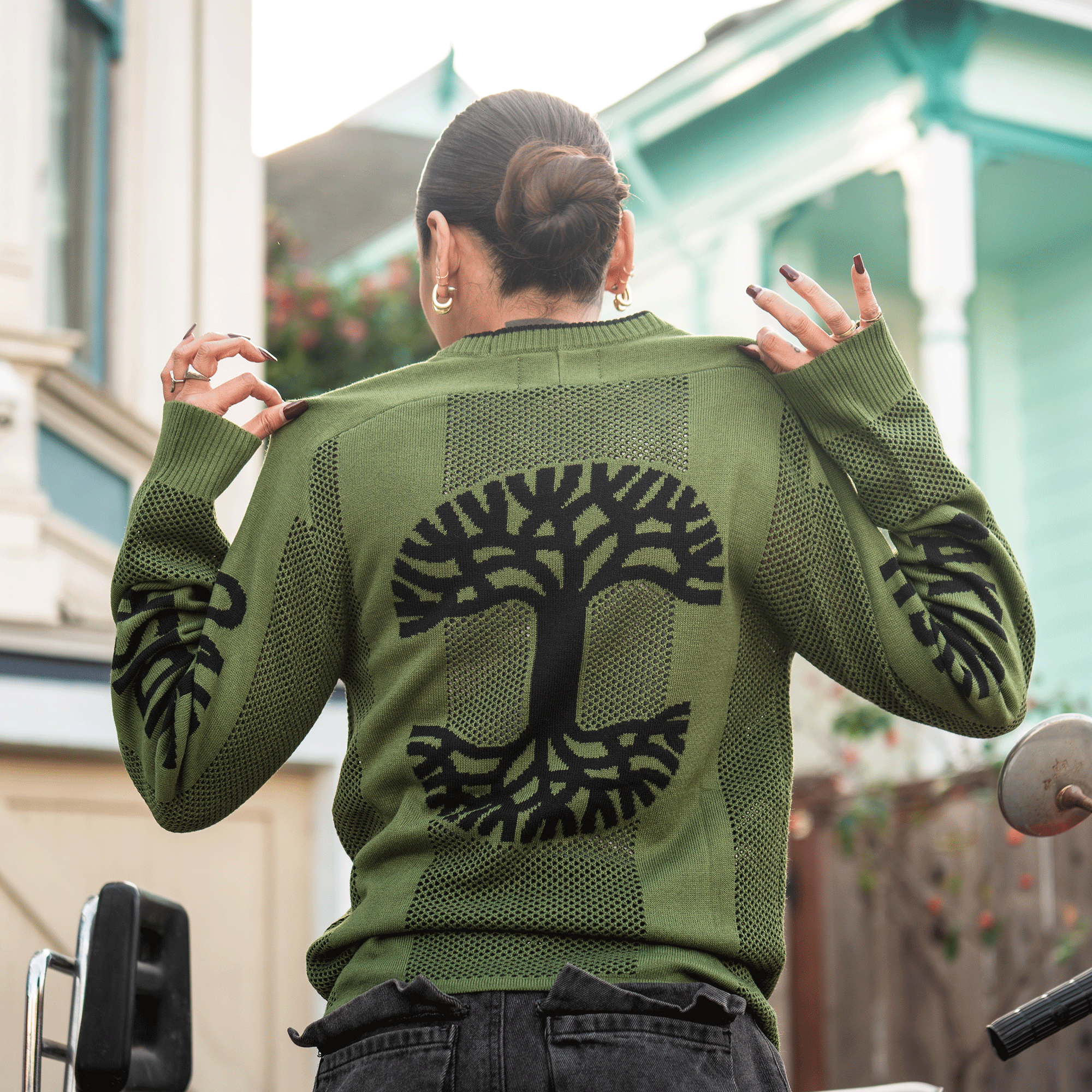 A person with dark hair in a bun wears the Oaklandish Terrace Sweater by Oaklandish, featuring an army green knit and bold black tree design on the back and sleeves. They hold up the collar outdoors, accessorized with gold hoops and black nail polish.