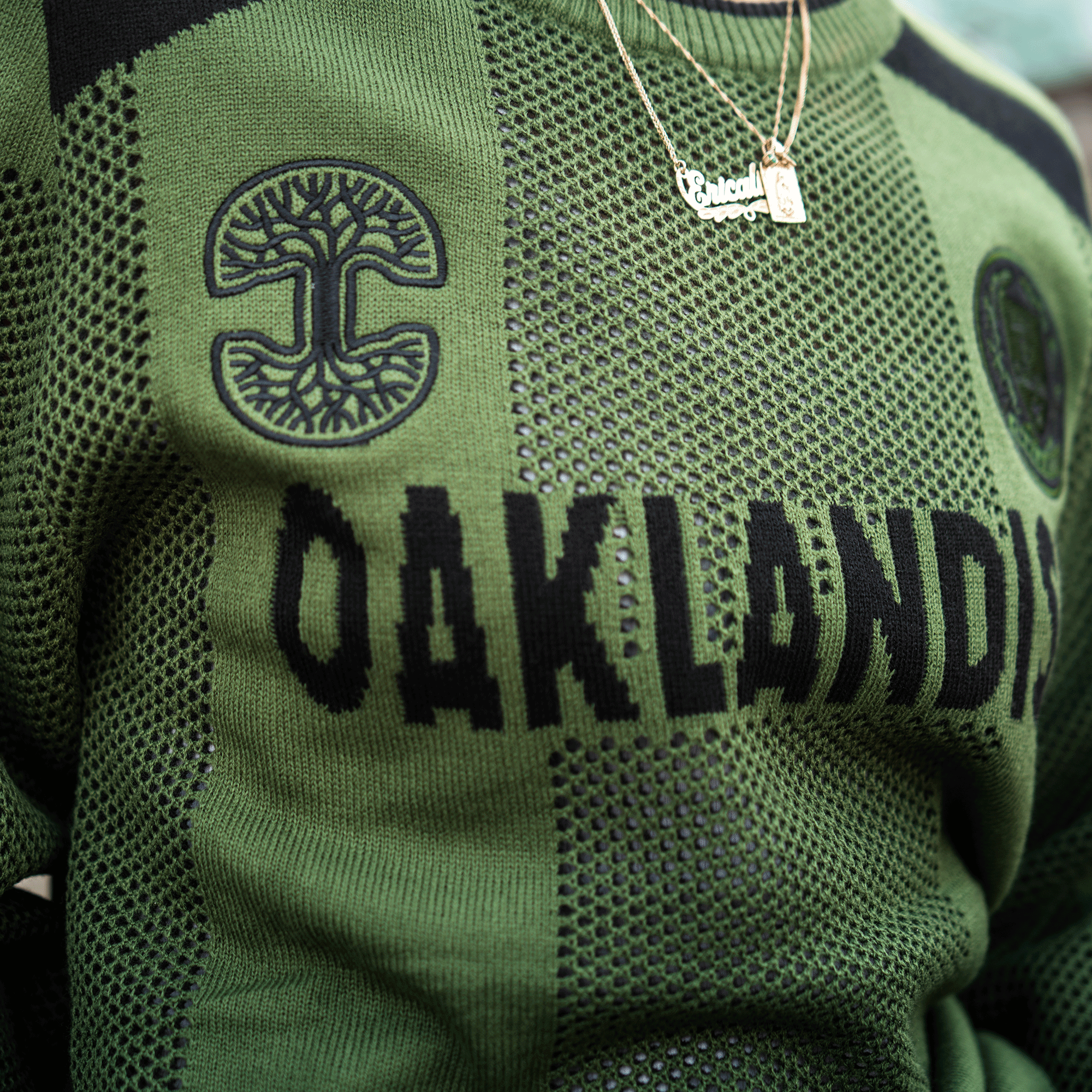 A close-up of the Oaklandish Terrace Sweater—a green knit unisex piece with “OAKLANDISH” and a black tree symbol—styled with two gold necklaces: one nameplate pendant and one rectangular charm. Brand: Oaklandish.