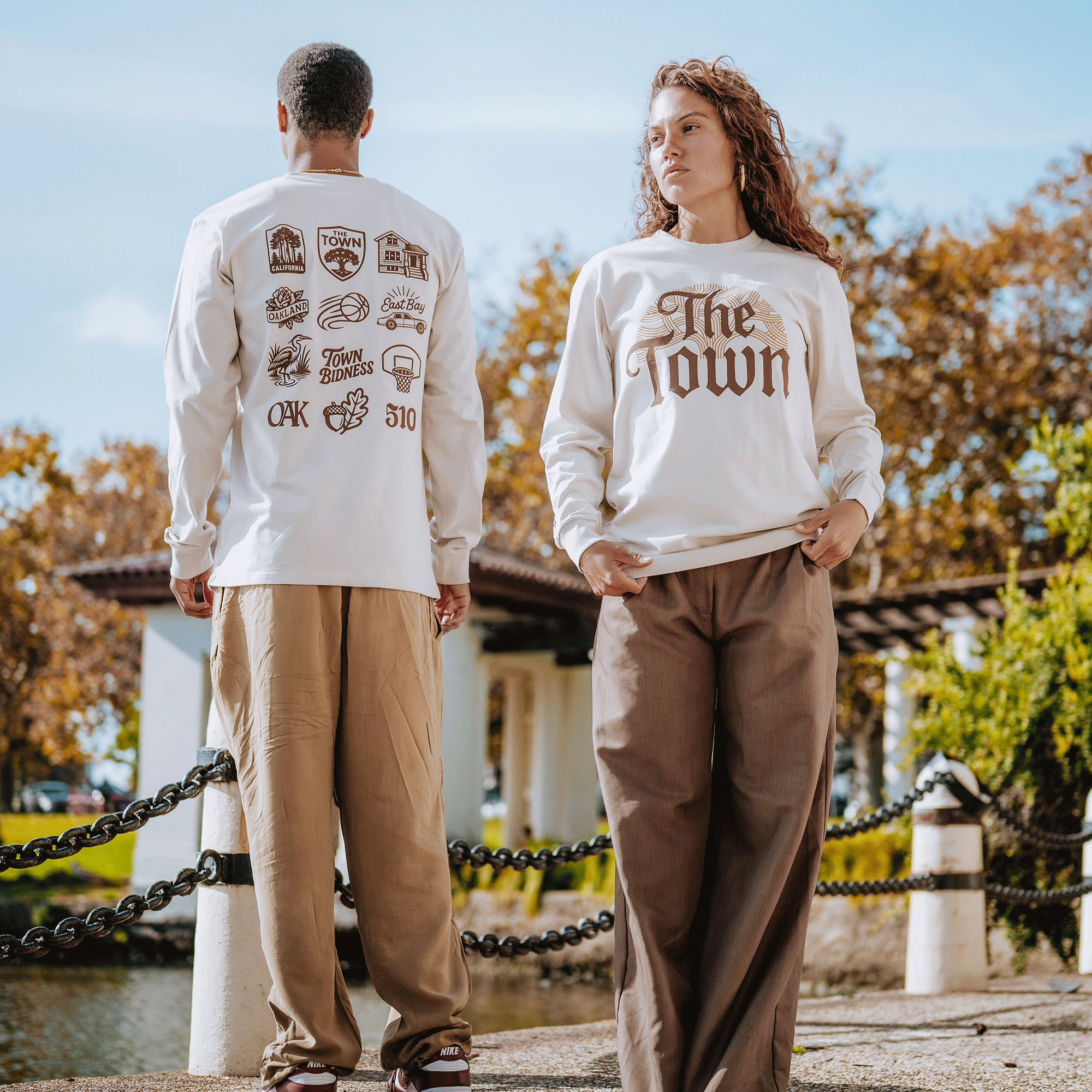 Two people model GSW's Pirate Black-Letter LS Tee outdoors. The woman, facing forward, wears the tee with bold script—a tribute to Golden State Warriors pride—set against fall trees and a pavilion background.