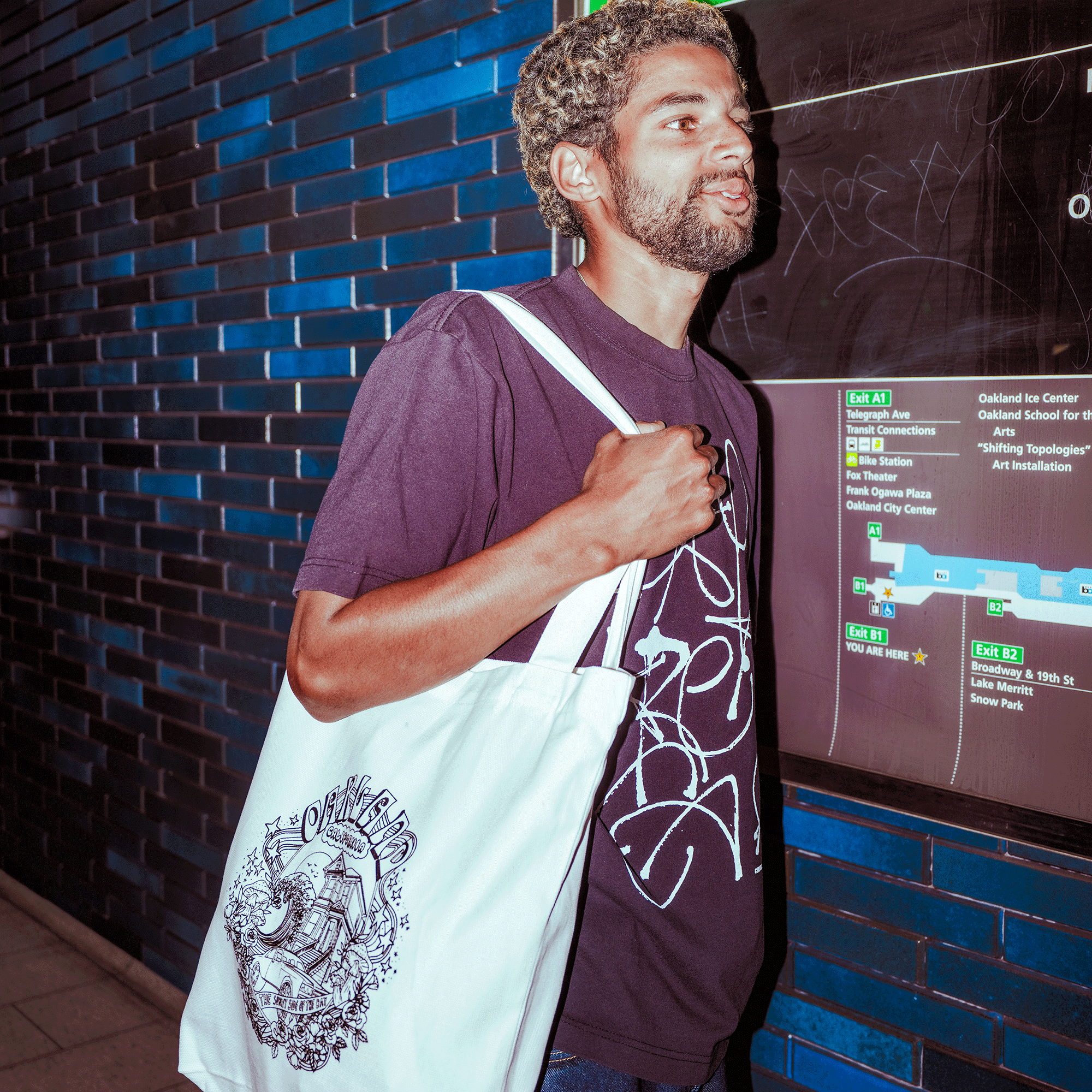 A man with curly hair and a beard, wearing an Oaklandish Presto Tee—men's black t-shirt with white abstract designs in a classic fit—stands near a green and black transit map, carrying a heavy-weight cotton tote bag with detailed black artwork.