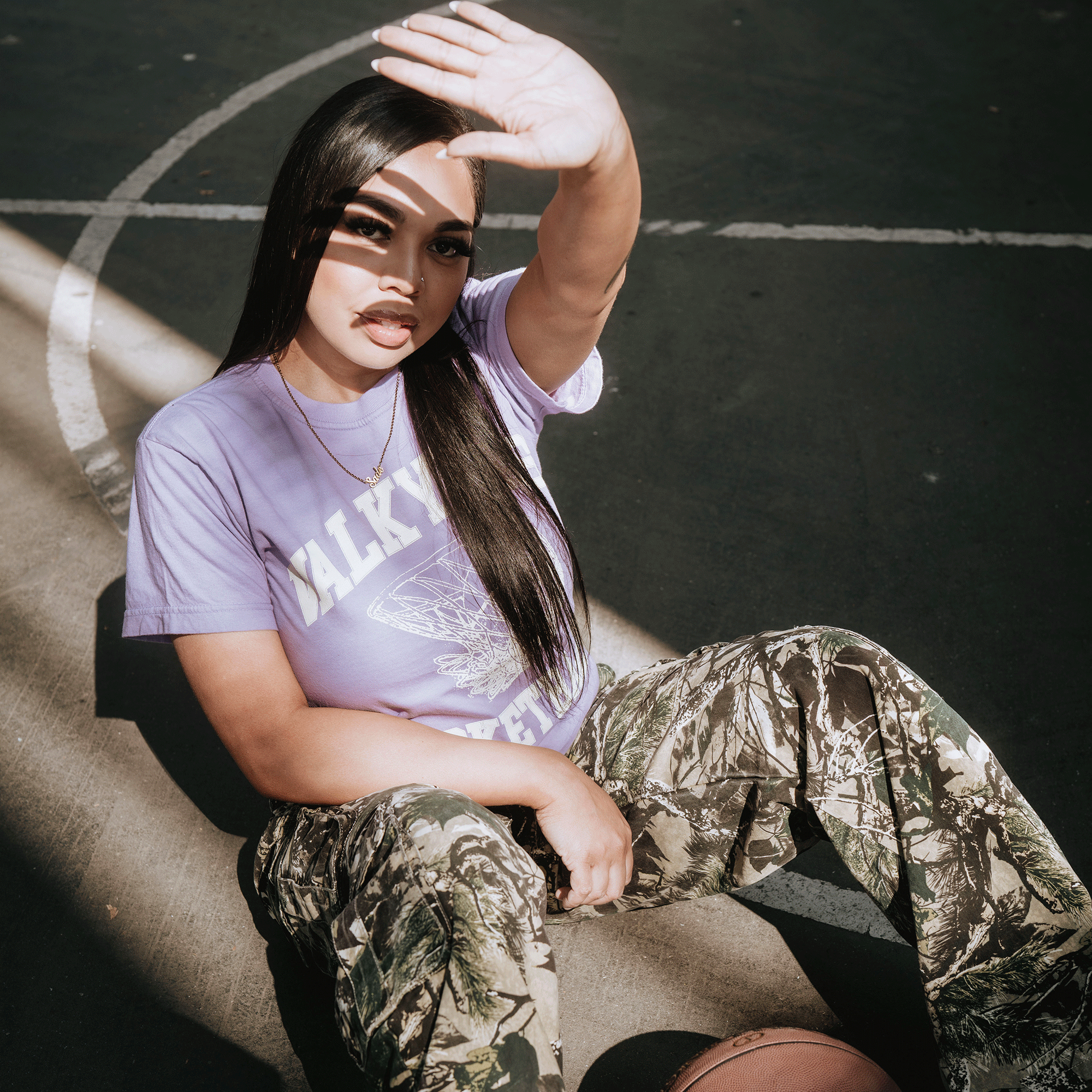 A young woman sits on an outdoor basketball court, shielding her eyes from the sun. She wears the GSV Golden State Valkyries Rec League Premium Tee and camouflage pants, sunlight casting strong shadows across her face.