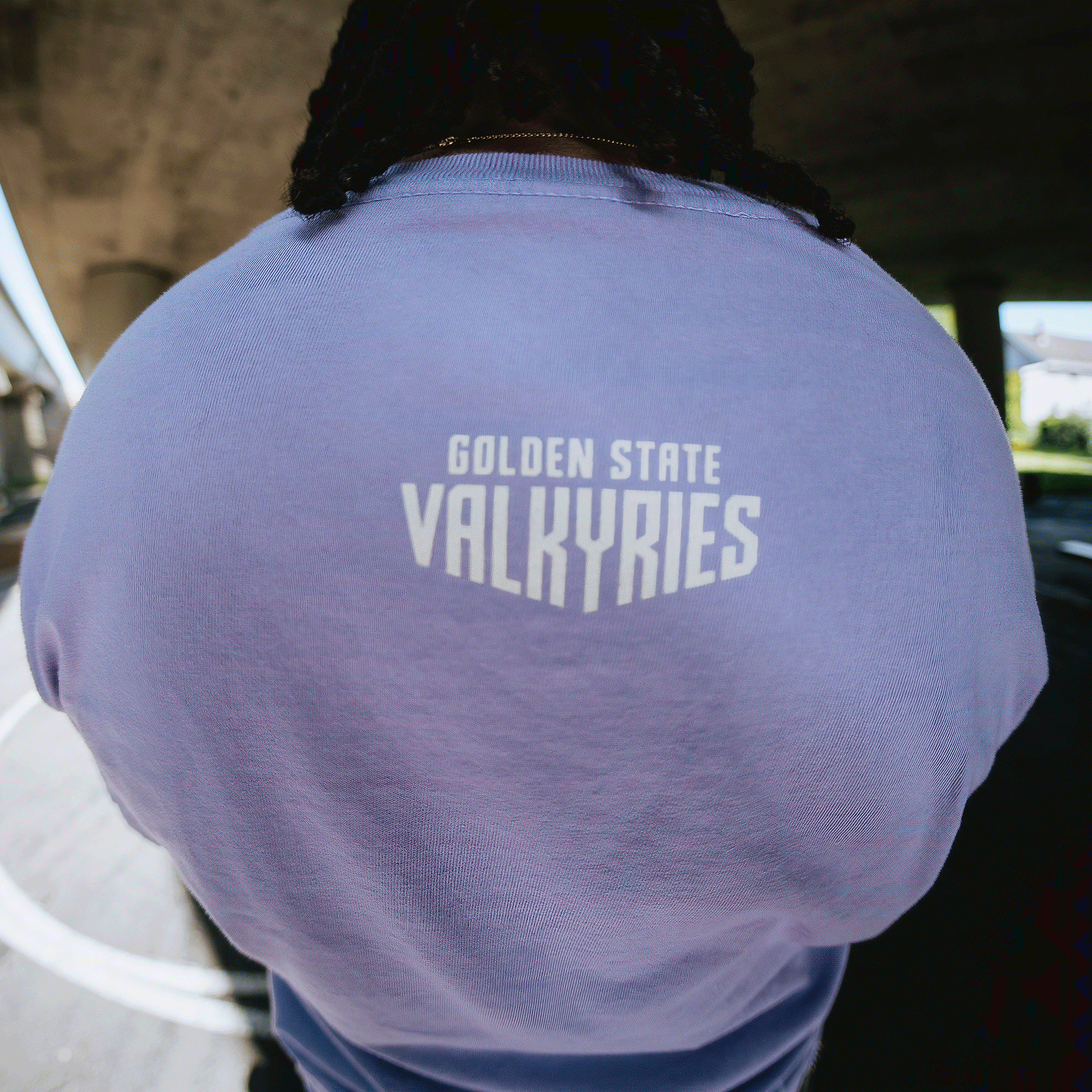 A person faces away from the camera, wearing the GSV Golden State Valkyries Rec League Premium Tee in light purple with “GOLDEN STATE VALKYRIES” printed in bold white on the upper back, standing outdoors beneath a bridge.